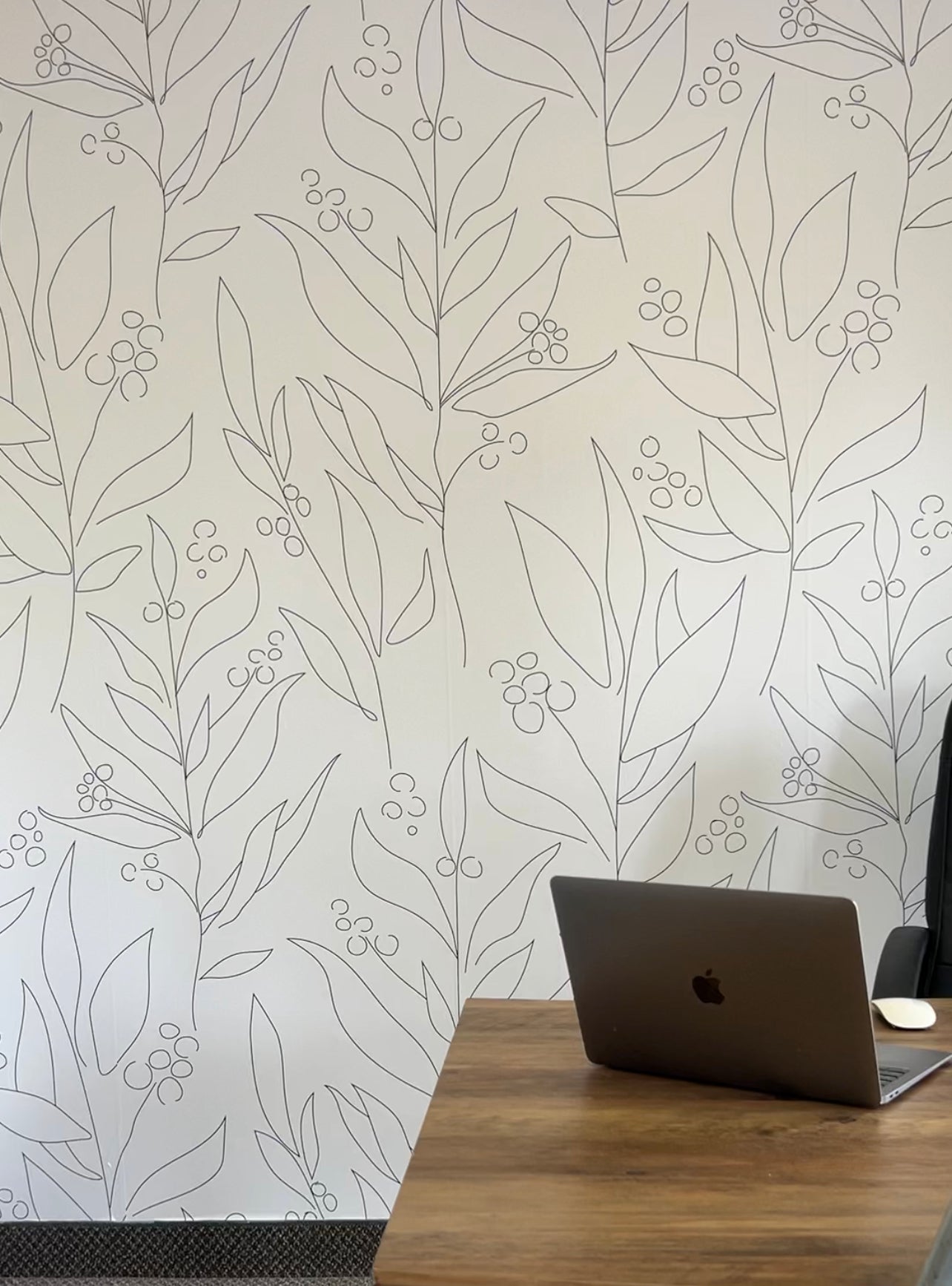 A modern office setup with a wooden desk, an open laptop, and a black office chair. The wall features white wallpaper with a delicate black abstract floral pattern, creating a stylish and professional atmosphere.