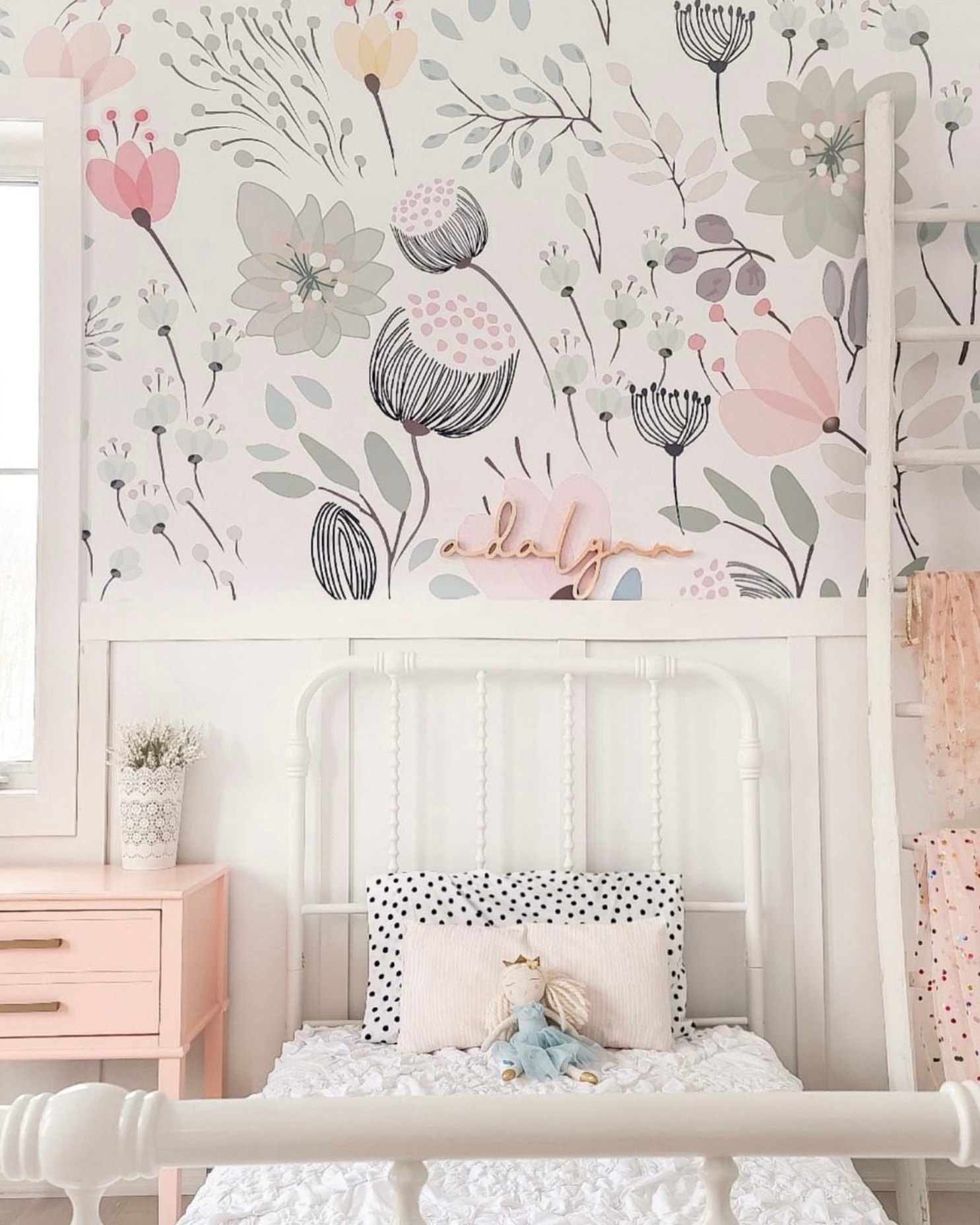 A cozy corner of a child's bedroom with the pastel floral wallpaper providing a delicate backdrop to a white metal bed adorned with a pink bedspread, a white and polka dot pillow, and soft toys, creating a dreamy and comforting atmosphere