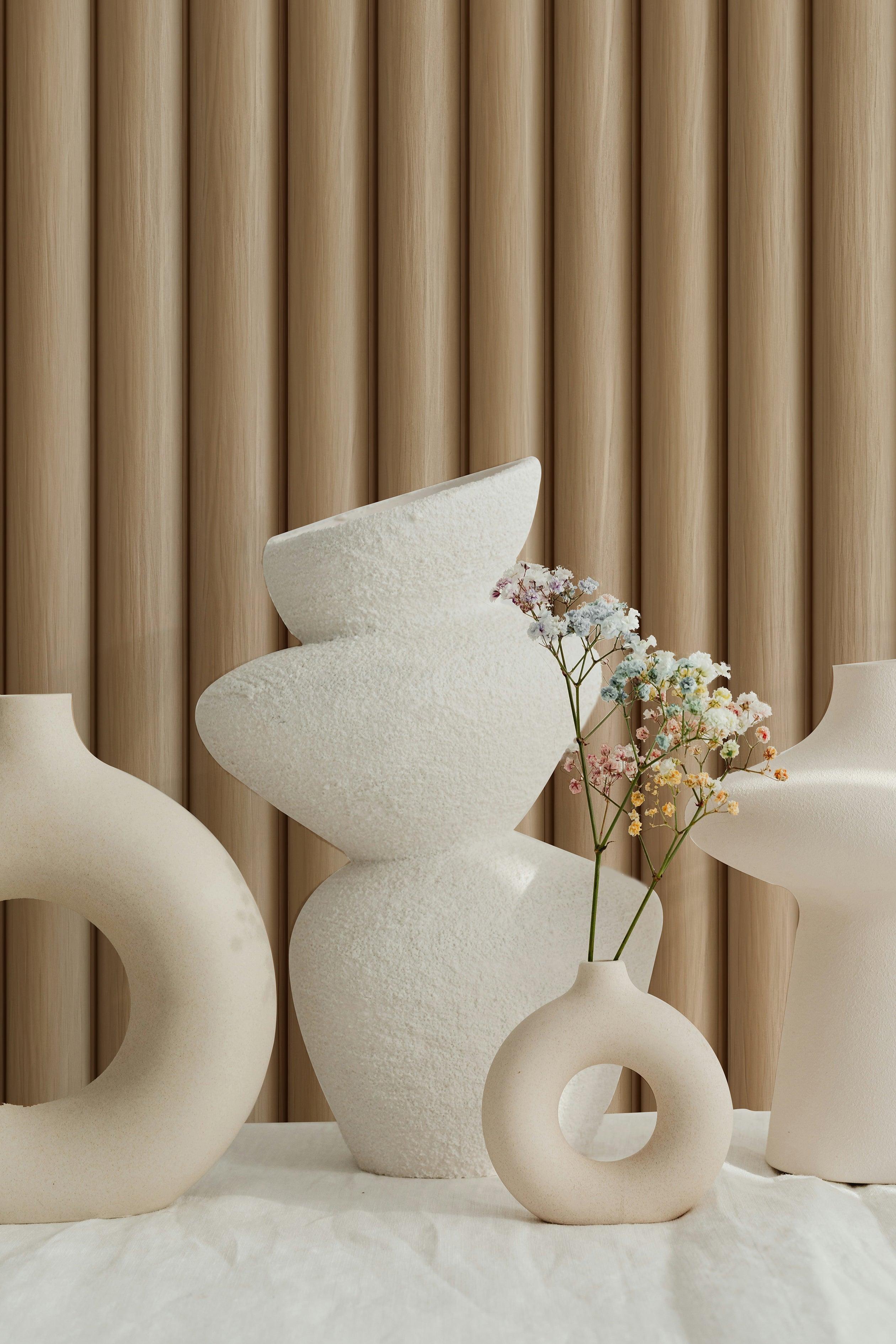 An arrangement of modern ceramic vases in various shapes and textures, set against the backdrop of Wooden Pillar Wallpaper with vertical grooves in a warm wood tone. The wallpaper's texture enhances the visual appeal of the minimalist decor.