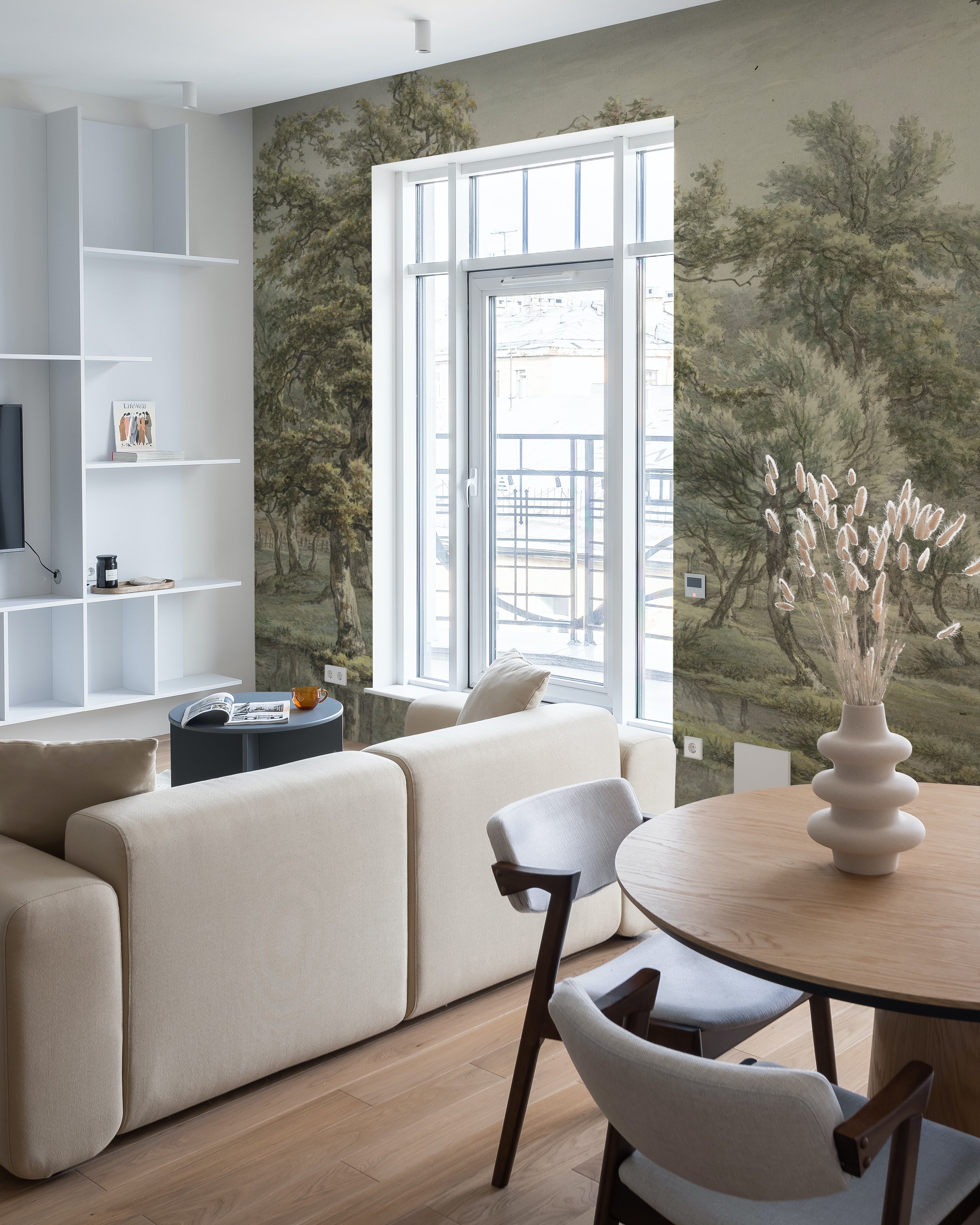 A bright and airy living room corner with a comfortable beige sofa and wooden chairs, looking out to a balcony. The wall is covered by the 'Landscape in Eext' mural, providing a stunning backdrop of vintage greenery and tranquil nature.