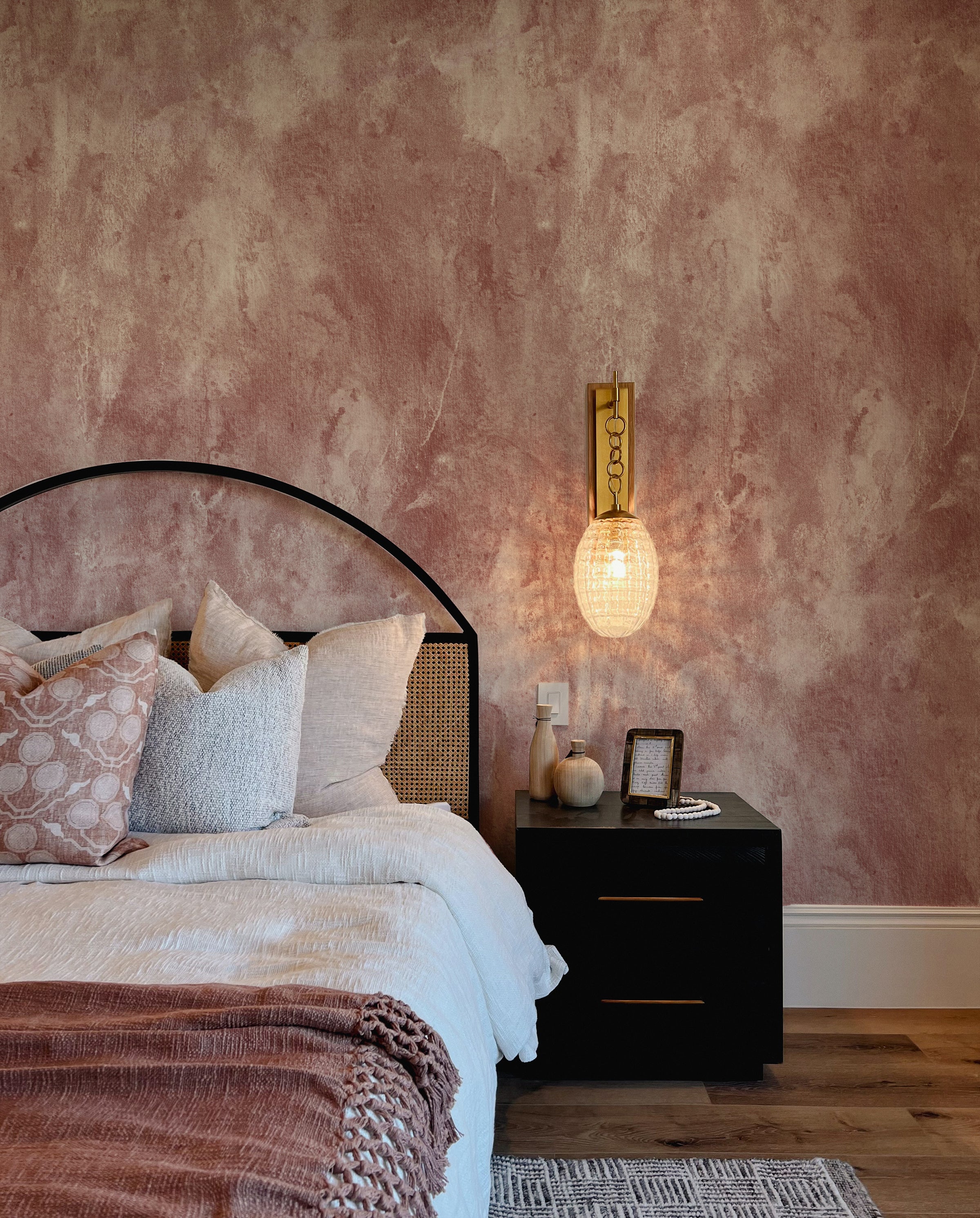 A stylish bedroom with a contemporary vibe, showcasing a black metal bed frame dressed in neutral linens with dusty rose accents. The Dusty Rose Limewash Wallpaper envelops the room in a cozy, inviting atmosphere.