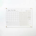 The full view of the same acrylic board, mounted on a white wall with gold pins. The board includes a monthly calendar, sections for goals, tasks, a quote of the day, a water tracker, a menu, and notes. The design features a neutral color palette with small star illustrations, making it both functional and decorative.