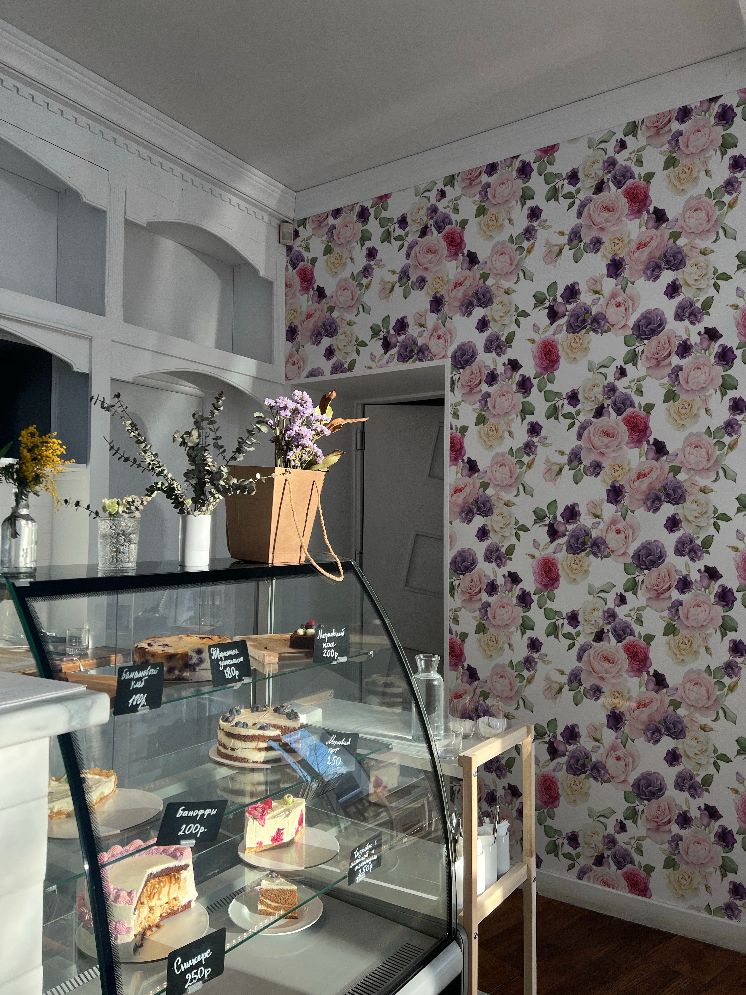 A charming cafe interior showcasing Mahalia Rose Floral Wallpaper, adorned with a lush pattern of pink and purple roses. The floral wall serves as a vivid backdrop to a display case filled with delectable cakes and pastries, creating a welcoming and stylish ambiance