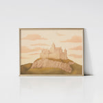 An art print of a whimsical pastel-colored castle framed in gold. The castle is set on a rocky hill with a backdrop of a dreamy sky filled with pink clouds, creating a magical and enchanting scene.