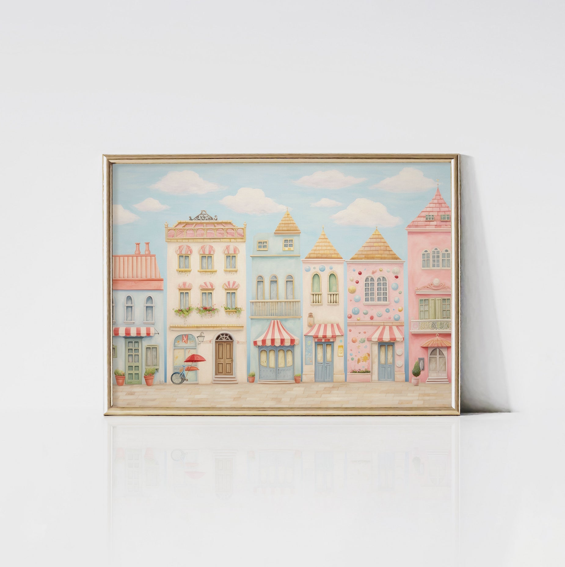 An art print of whimsical pastel-colored townhouses framed in gold. The buildings display different architectural styles, complete with awnings, balconies, and decorative elements, set against a clear blue sky with fluffy white clouds.