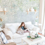 A cozy living space highlighted by the cherry blossom canopy wallpaper, with a child engrossed in a book on a white sofa. The mural's ethereal blossoms create an enchanting background that's both playful and serene.
