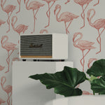 A stylish white speaker from Marshall is placed atop a small white cabinet against a backdrop of the Flamingo Party Wallpaper, which features a playful pattern of pink flamingos on a white background. The scene is complemented by a large green leafy plant in the foreground, adding a touch of nature to the decor.