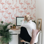 A modern home office setup against a wall adorned with Flamingo Party Wallpaper. A woman, seen from behind, is seated at a white desk with a computer, surrounded by green plants and soft pink accessories that echo the playful and vibrant theme of the flamingo pattern on the wall.