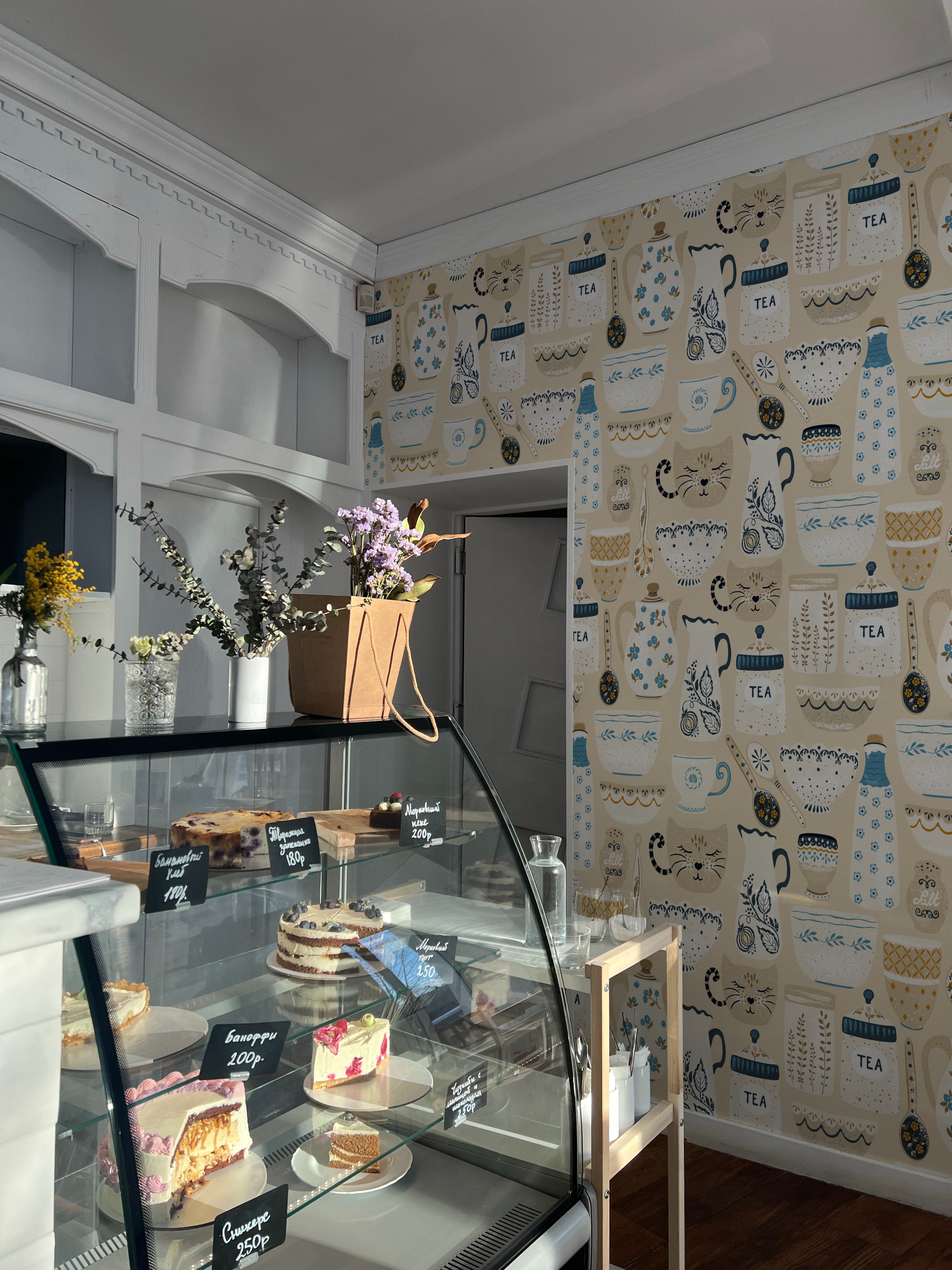 A cozy interior of a cafe with stylish decor, featuring walls adorned with 'Spring Ceramics Wallpaper'. The wallpaper displays charming illustrations of teapots, teacups, and other ceramics in soft blue, beige, and yellow tones. A glass display counter showcases a variety of appealing cakes and desserts, enhancing the warm and inviting atmosphere.