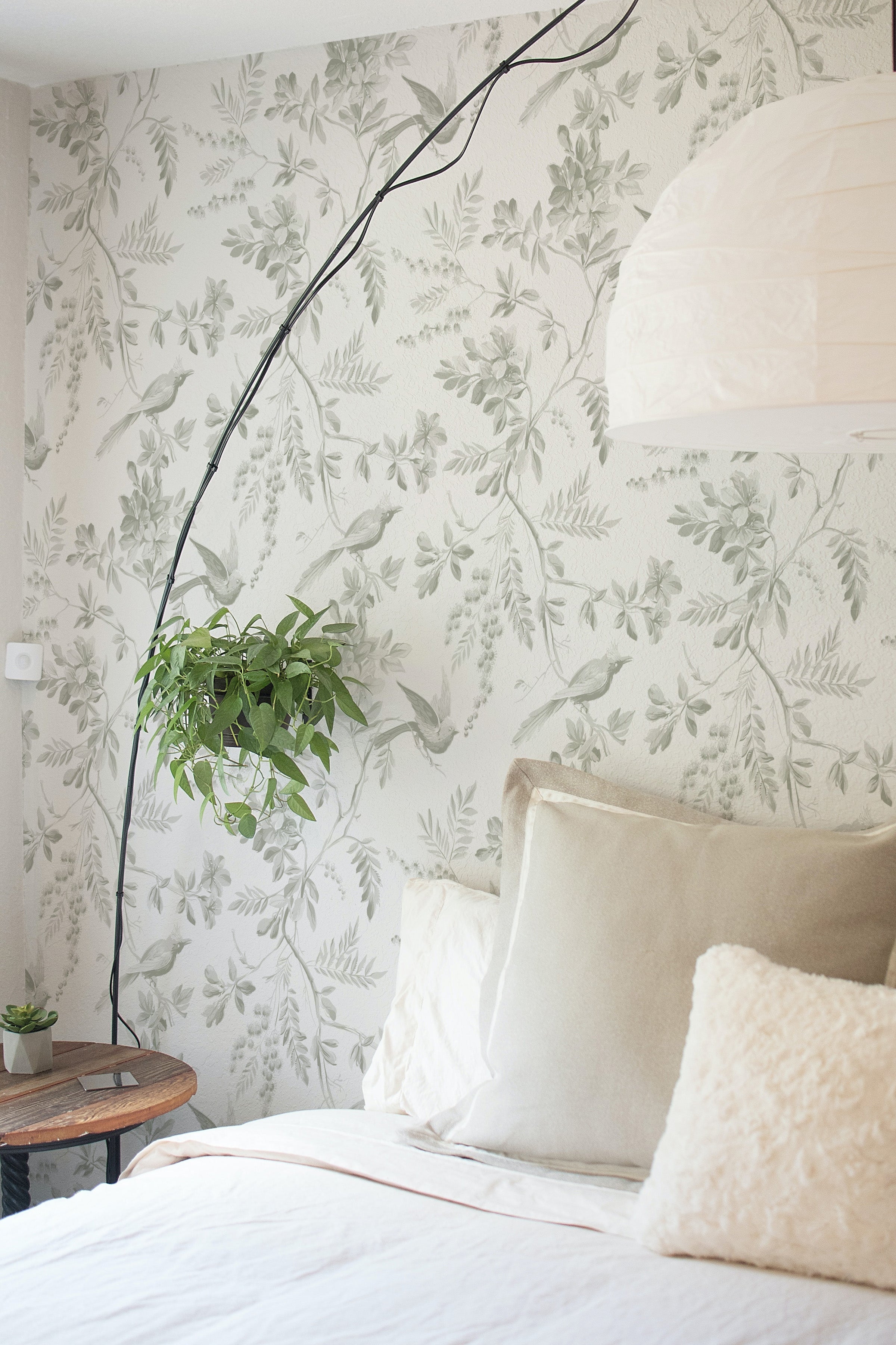 A bedroom scene with 'Blossoming Birds Wallpaper' creating a tranquil ambience, accented by an overhanging plant and a bed dressed in neutral linens, evoking a sense of being in a serene, bird-filled garden