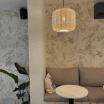 A cozy corner of a café with comfortable seating against a backdrop of 'Blossoming Birds Wallpaper', displaying a serene pattern of greenery and birds, complemented by a woven light fixture.