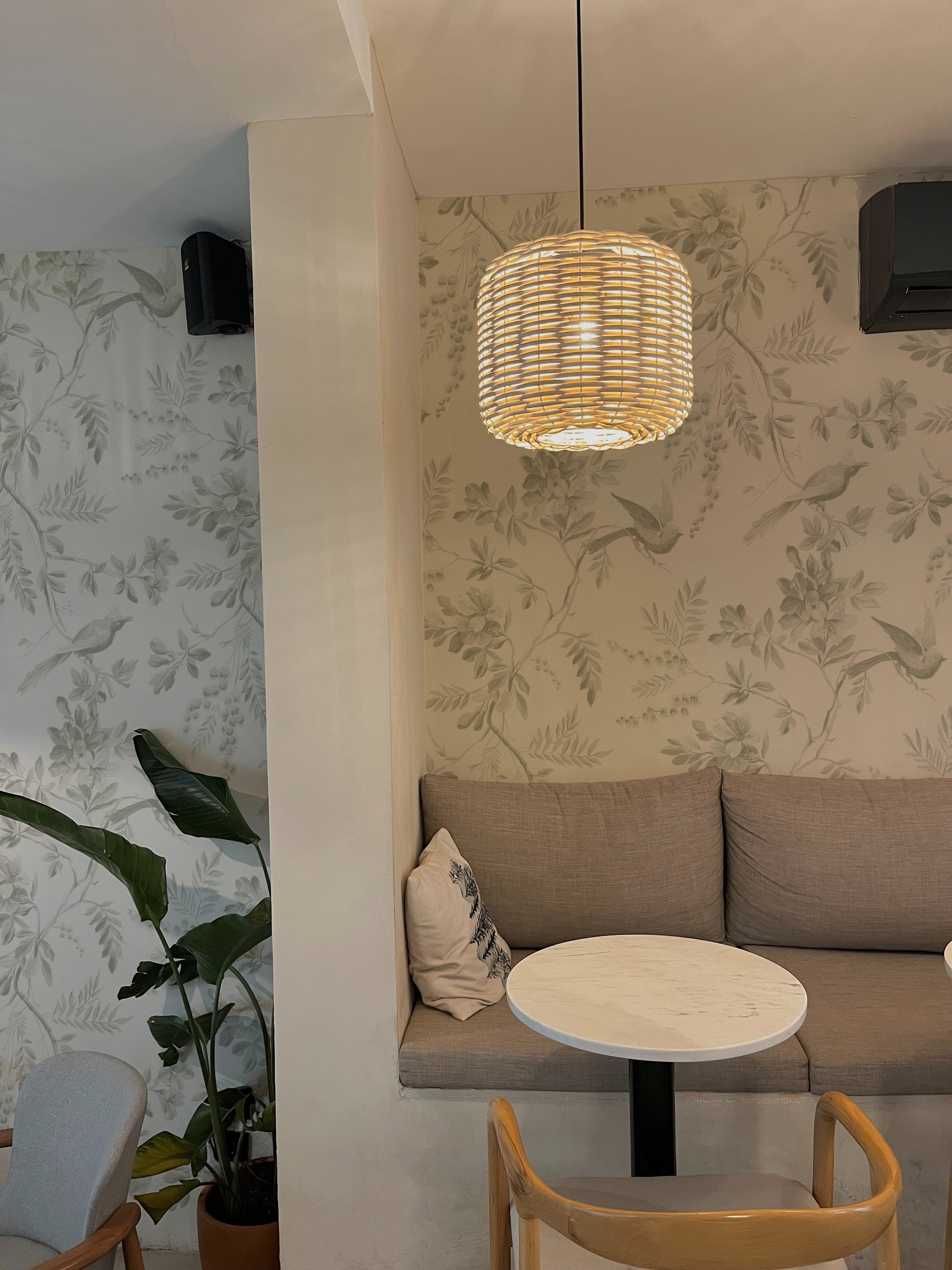 A cozy corner of a café with comfortable seating against a backdrop of 'Blossoming Birds Wallpaper', displaying a serene pattern of greenery and birds, complemented by a woven light fixture.
