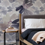 A cozy bedroom interior decorated with Purple Passion Wallpaper that displays an artistic arrangement of large floral patterns in muted purple and grey tones. The soft, expressive backdrop complements the simple wooden bed frame and dark bedding, adding a sophisticated and tranquil feel to the room