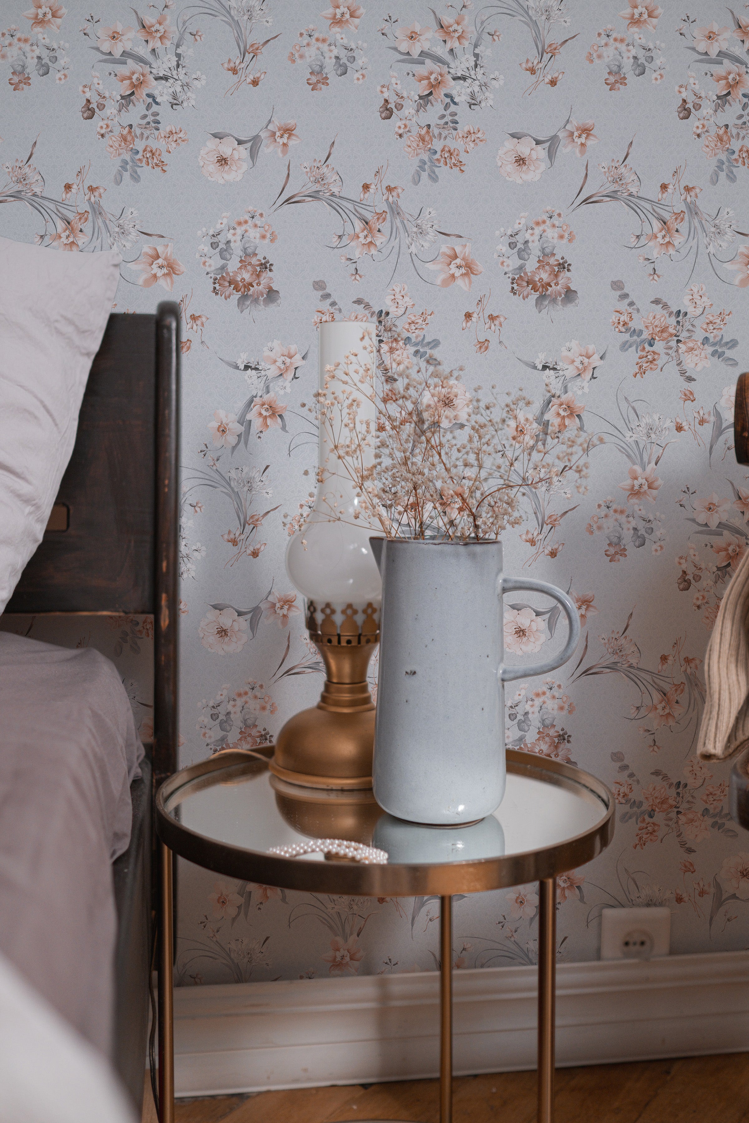 A charming bedroom setup highlighting the Boudoir Blooms Wallpaper which serves as a backdrop for a nightstand scene. The wallpaper's floral pattern in peach and gray complements the vintage brass lamp and ceramic vase, adding a touch of elegance to the intimate space