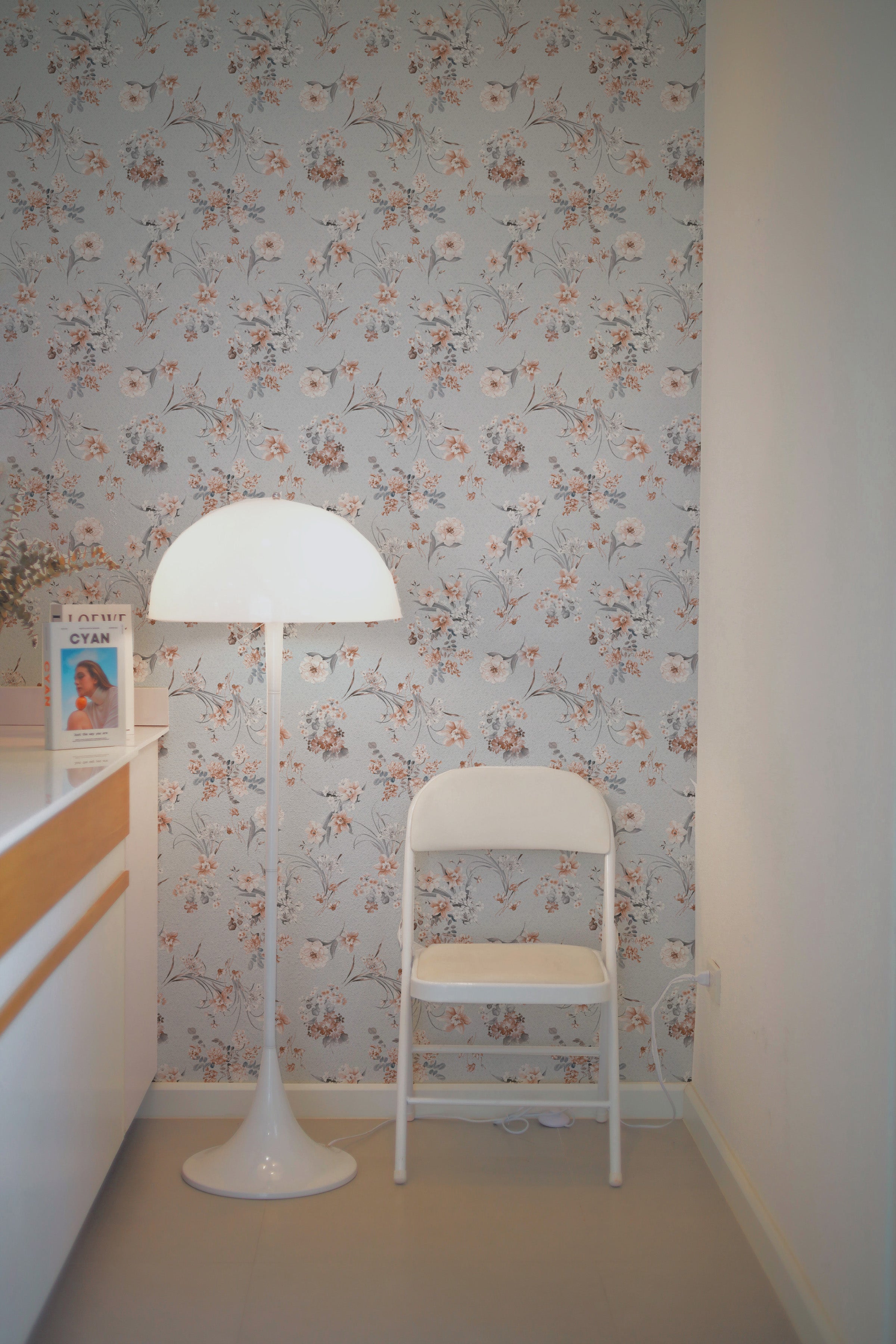 A cozy corner in a modern room featuring the Boudoir Blooms Wallpaper, adorned with a pattern of delicate floral blooms in shades of peach and gray. A sleek white chair and a stylish floor lamp complement the wallpaper, creating a serene and inviting space.
