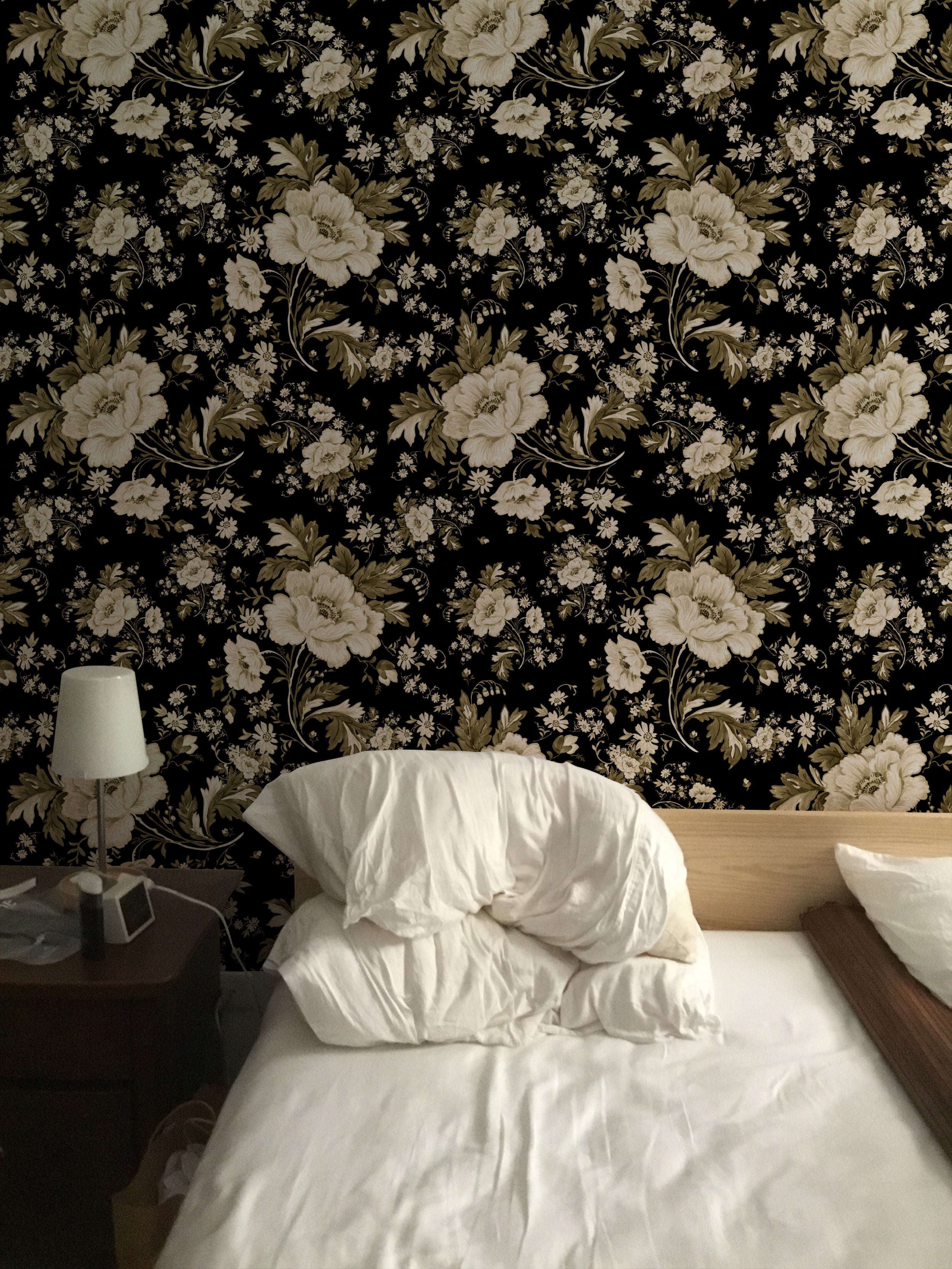 A cozy bedroom scene where the Midnight Garden Wallpaper creates a striking backdrop. The black background with white floral prints offers a bold contrast to the soft white bedding and the small bedside lamp, crafting a tranquil yet sophisticated sleeping area.