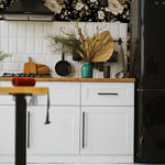 A modern kitchen scene featuring Midnight Garden Wallpaper, with its rich floral pattern of large white flowers on a dramatic black background. The space is stylishly accented with contemporary white cabinetry, a black refrigerator, and decorative elements such as a woven plant pot and gold details.