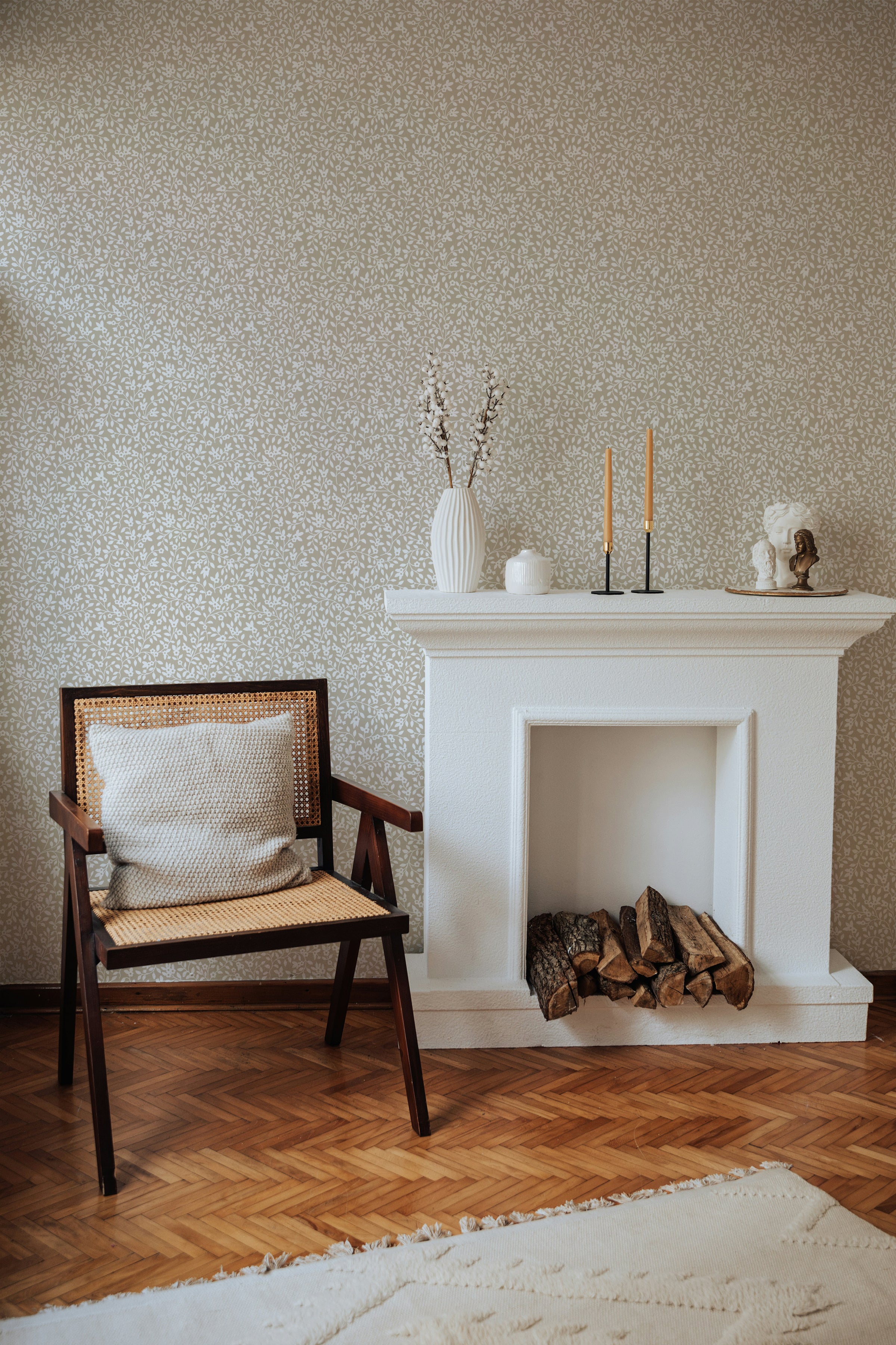 A warm and inviting room with herringbone wood flooring features a wall adorned with 'Floral Vines Wallpaper'. The wallpaper displays a dense pattern of taupe floral vines, providing a vintage charm. The decor includes a classic white fireplace with logs, a mid-century modern chair with a rattan back, and decorative objects atop the mantel.