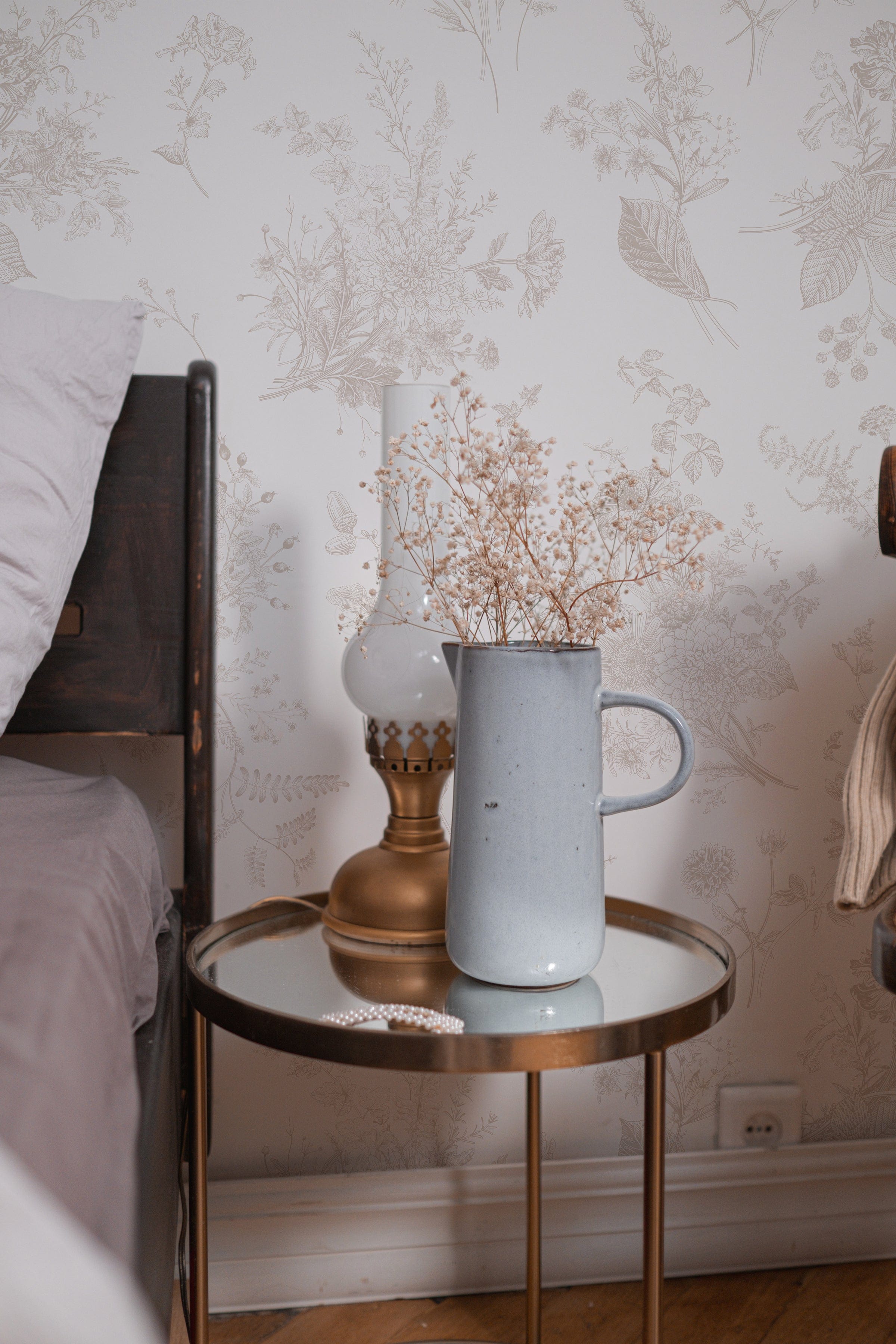 A cozy bedside scene with the Jouyful Garden Wallpaper in the background. A vintage wooden bedside table holds a pale blue ceramic pitcher filled with sprigs of dried flowers, accentuating the wallpaper's romantic and pastoral print. The gentle pattern of the wallpaper, with its muted tones and fine details, complements the simplicity and rustic charm of the bedroom decor.