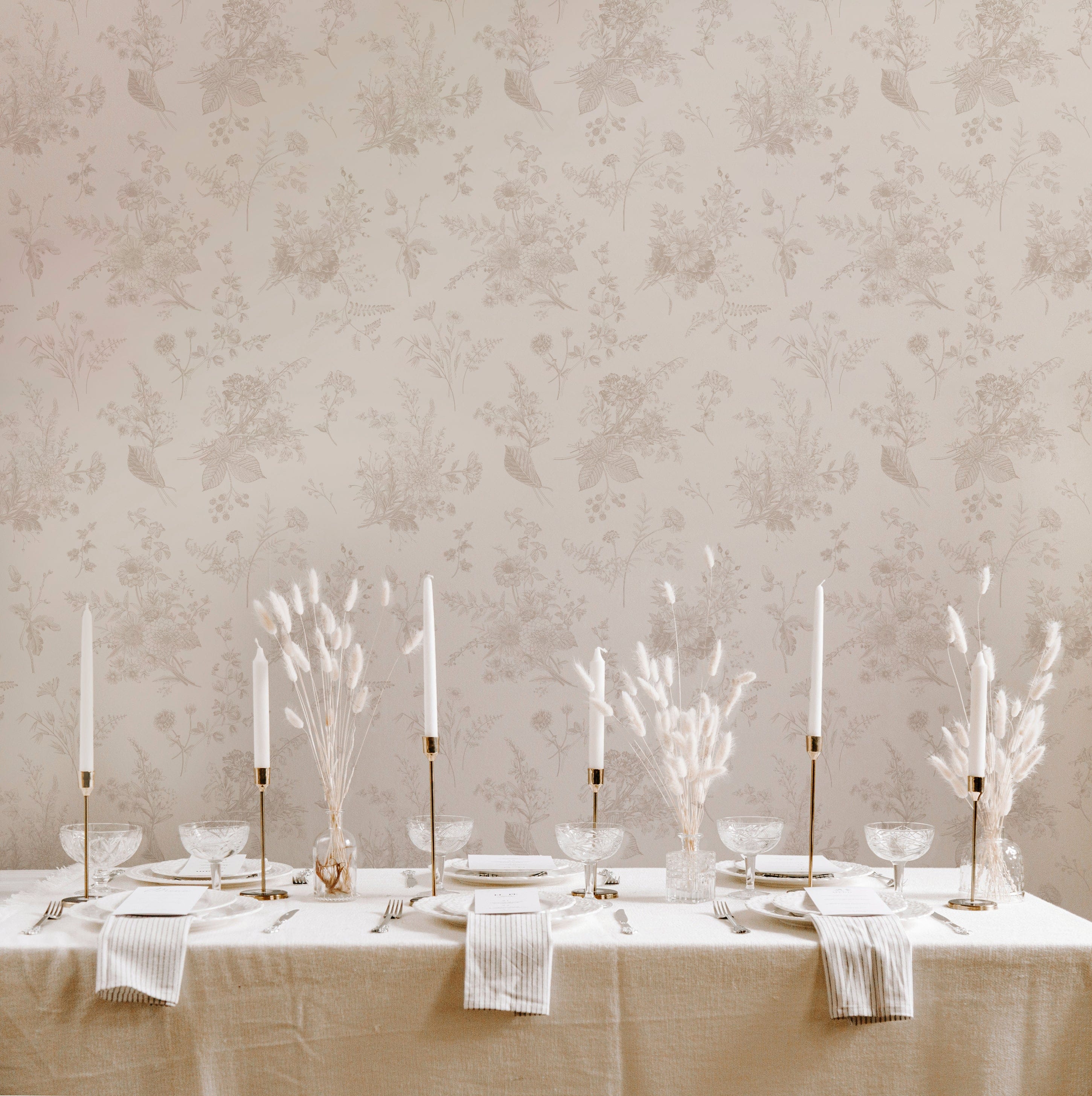 A dining area elegantly set against the Jouyful Garden Wallpaper, which features a delicate and traditional pattern of botanical sketches. The wall creates a refined and soft backdrop for the dining table, which is dressed in a natural linen tablecloth and set with white plates, clear glassware, and tall, slender candlesticks. The overall effect is one of understated luxury and pastoral charm.