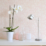 A section of 'Flower Power' wallpaper in a soft champagne color with white floral patterns, applied to a wall behind a shelf with a potted orchid, clear glass bottles, and candles