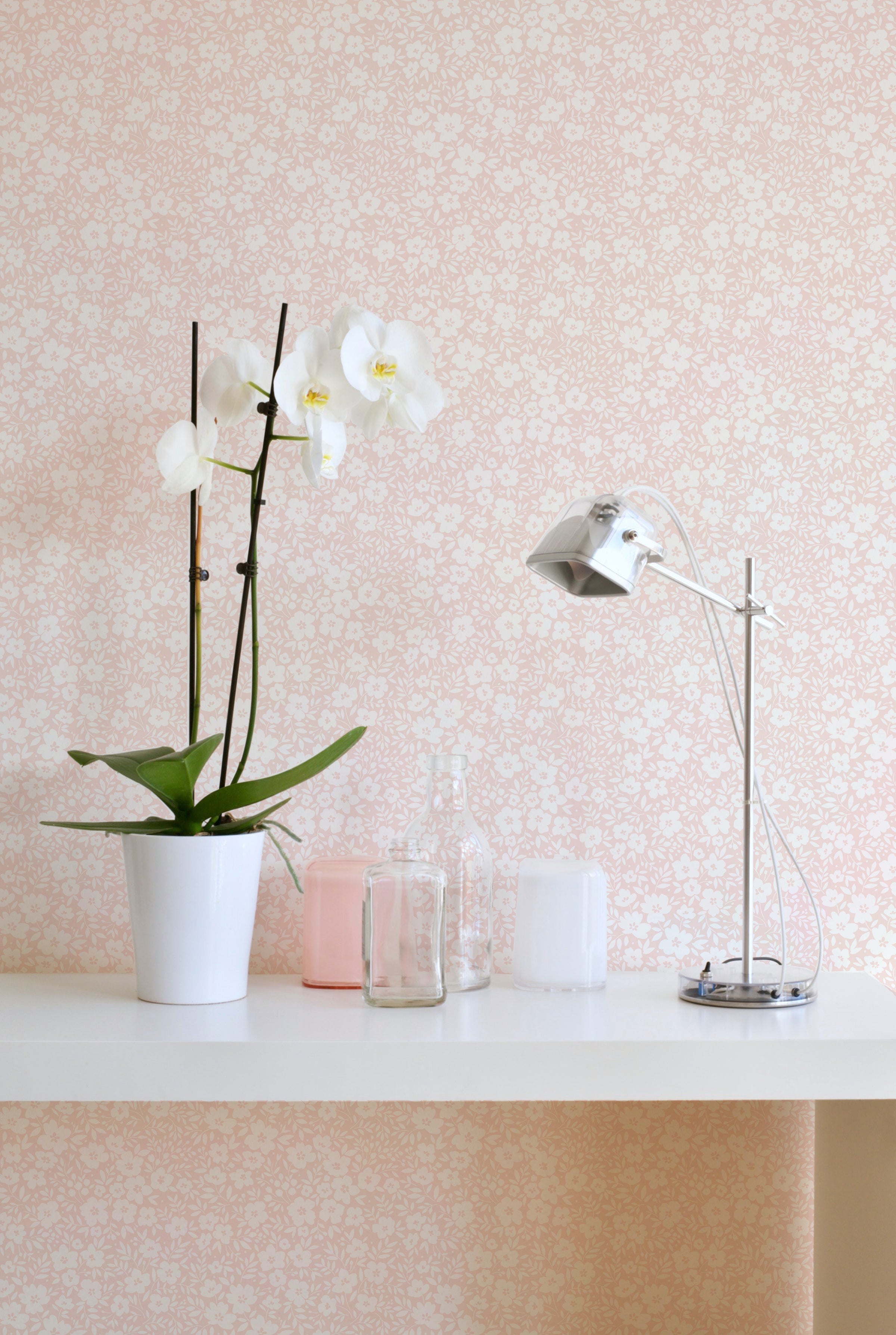 A section of 'Flower Power' wallpaper in a soft champagne color with white floral patterns, applied to a wall behind a shelf with a potted orchid, clear glass bottles, and candles