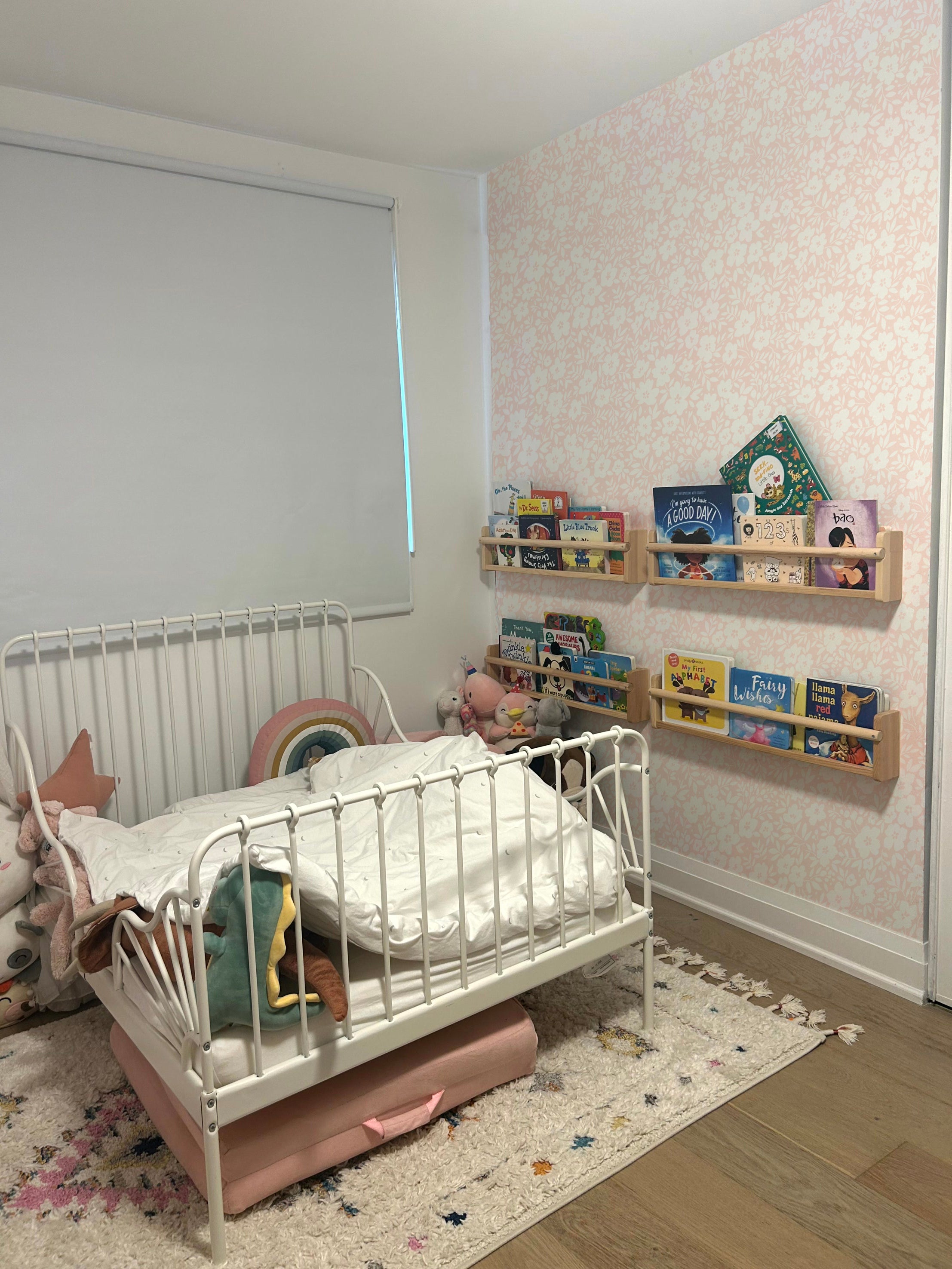 A child's bedroom featuring the Flower Power Wallpaper in a delicate pink floral pattern. The room includes a white metal bed with colorful bedding, surrounded by toys and children's books, creating a playful and inviting atmosphere.