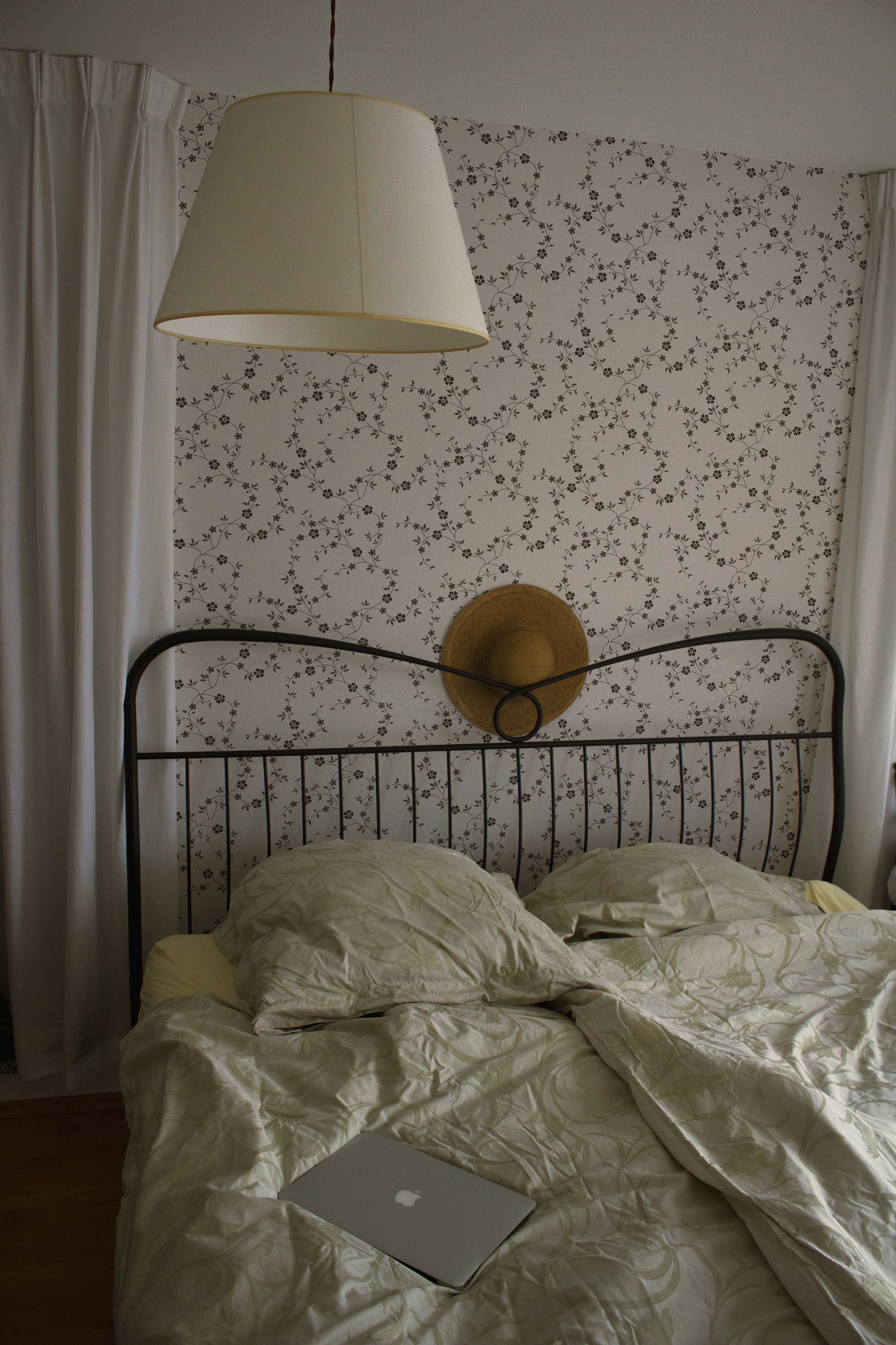 An intimate sleeping area adorned with Charming Floral Wallpaper in Deep Brown. The gentle floral motifs create a peaceful and serene environment, complemented by a simple black metal bed frame and crisp white bedding.