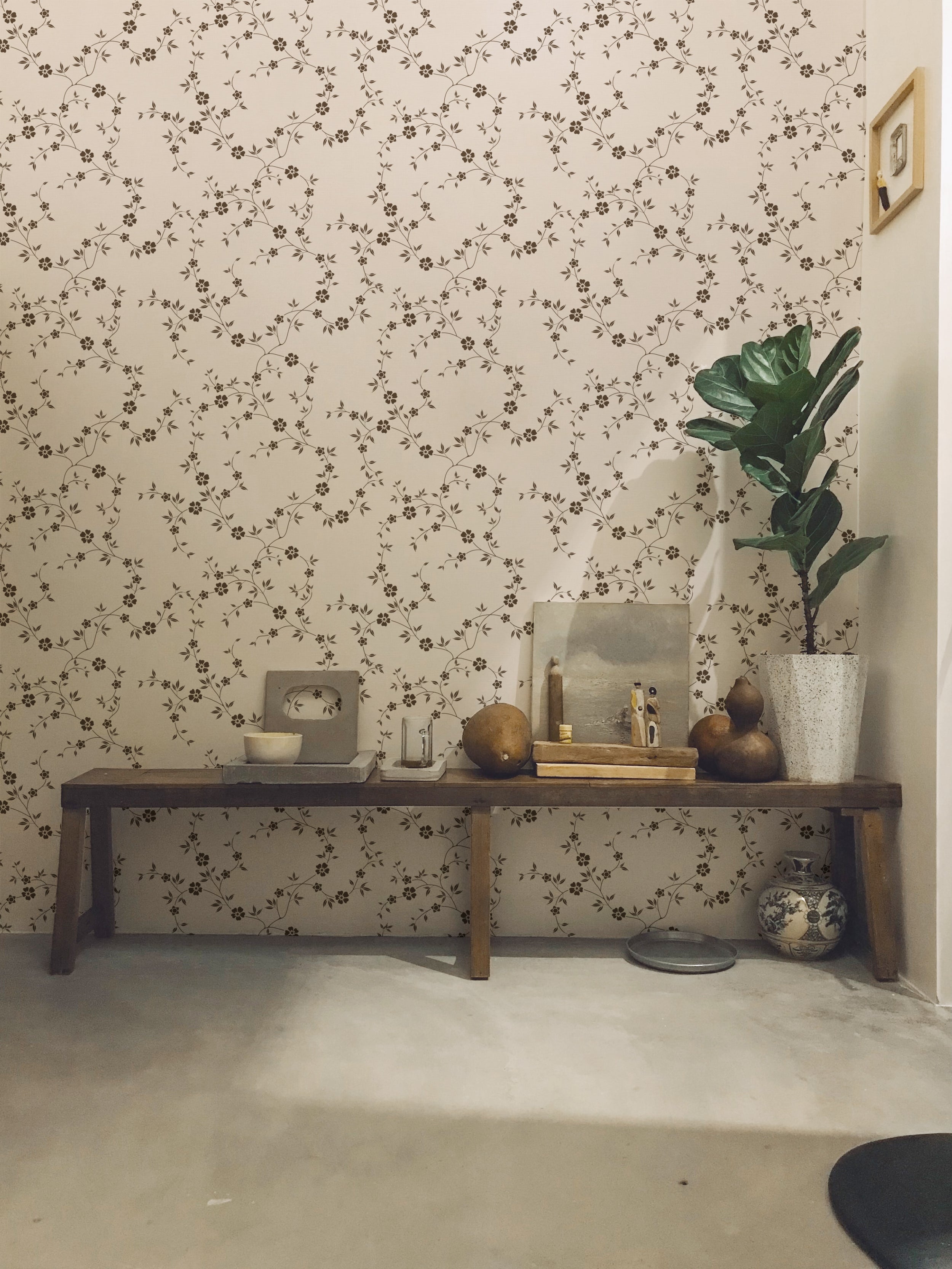 A sophisticated interior featuring Charming Floral Wallpaper in Deep Brown, showcasing an intricate pattern of small flowers and leaves. The setting includes a rustic wooden bench with decorative items, creating a warm, inviting ambiance.