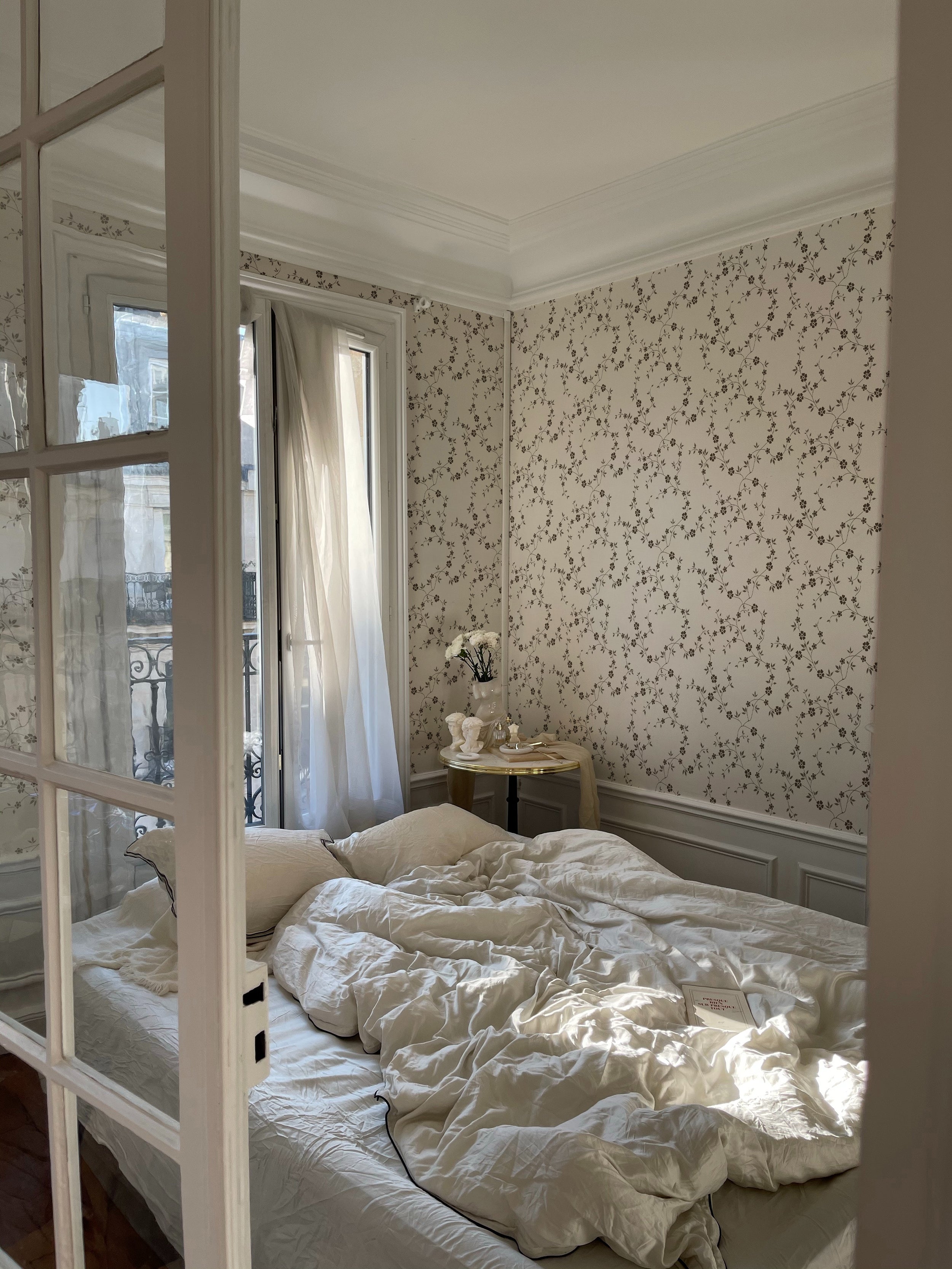 A cozy bedroom scene where the Charming Floral Wallpaper in Deep Brown provides a delicate backdrop. The natural light from a window illuminates the floral pattern, enhancing the room's vintage charm, complete with a classic bed and soft, inviting linens.