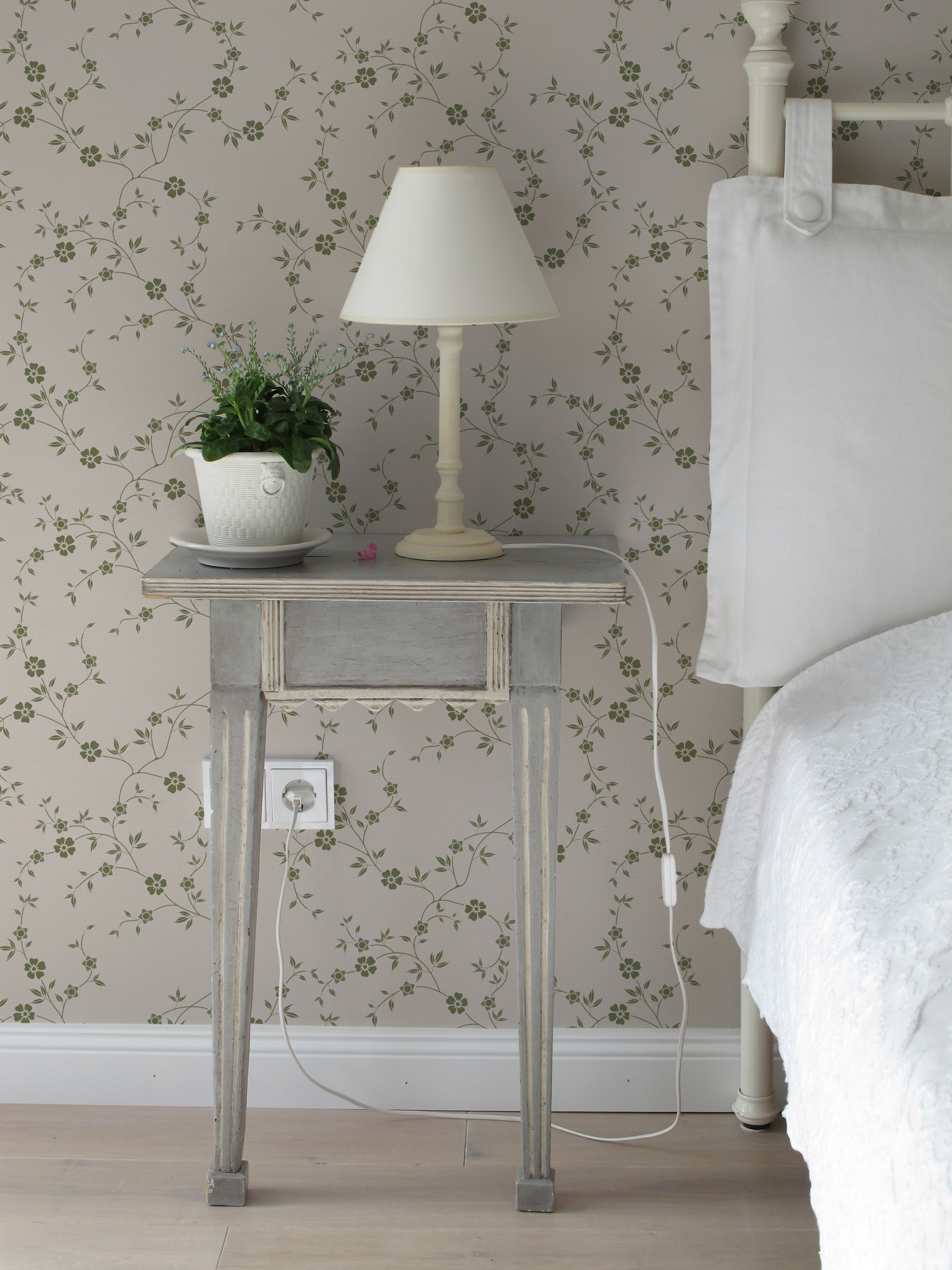 A cozy bedroom corner where the Charming Floral Wallpaper-Moss creates a relaxing ambiance. A distressed wooden side table holds a classic lamp and a potted green plant, complementing the natural theme alongside a comfortable bed with plush white bedding.