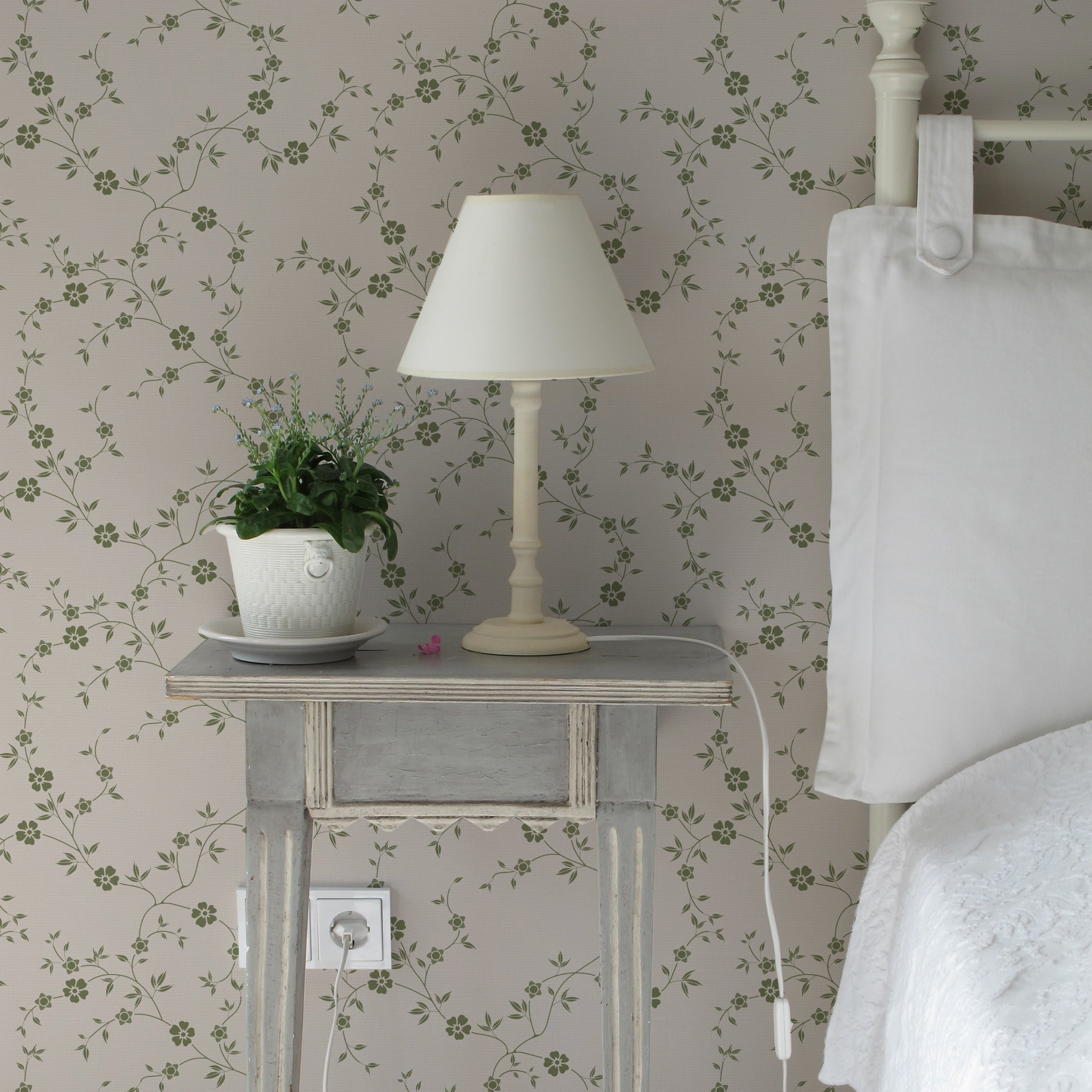 A cozy bedroom corner where the Charming Floral Wallpaper-Moss creates a relaxing ambiance. A distressed wooden side table holds a classic lamp and a potted green plant, complementing the natural theme alongside a comfortable bed with plush white bedding.