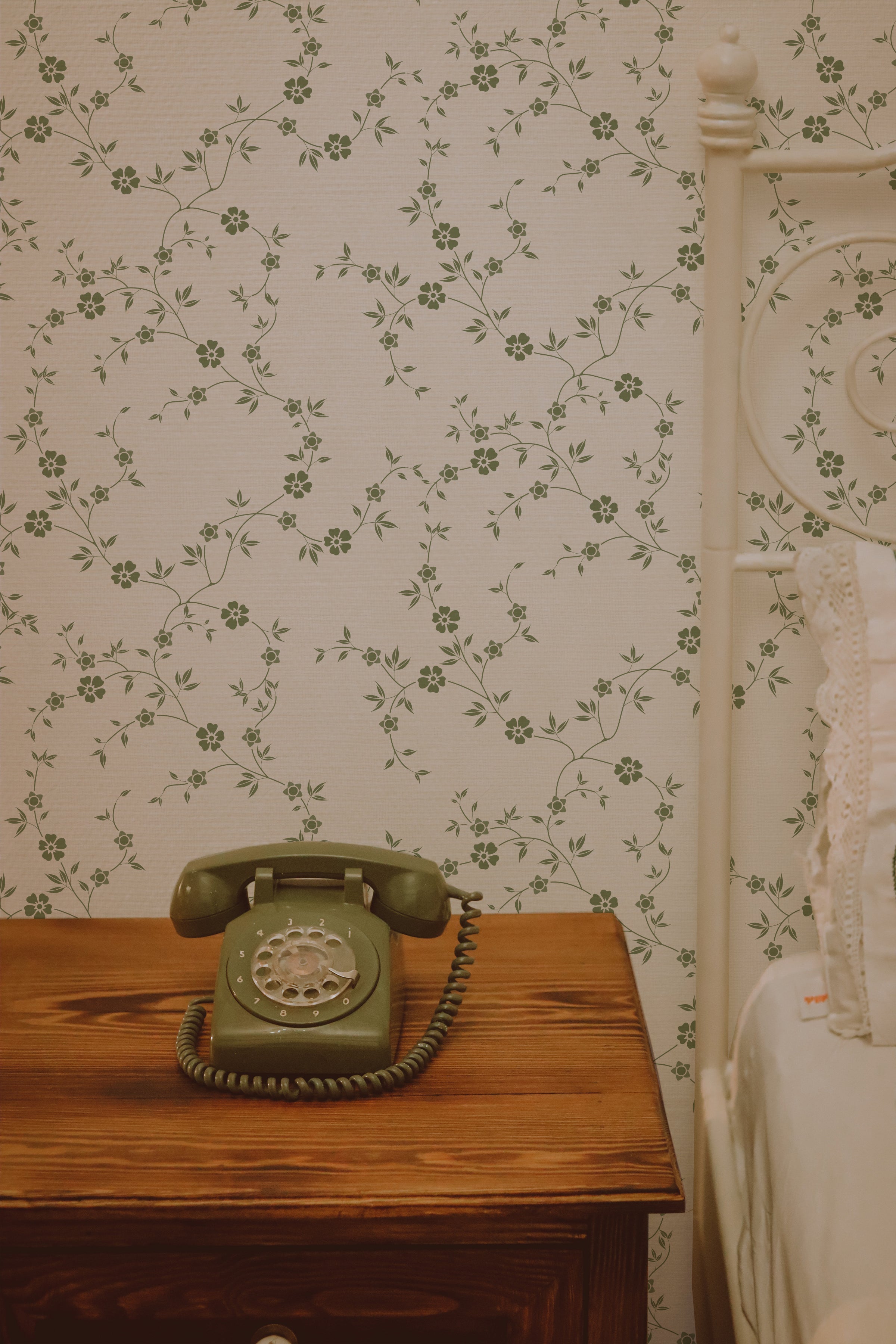 A vintage green rotary telephone sits on a wooden nightstand, next to an iron bed with white linens. The wall behind is adorned with the Charming Floral Wallpaper-Moss, featuring a delicate, intertwined green floral pattern on a warm beige background.