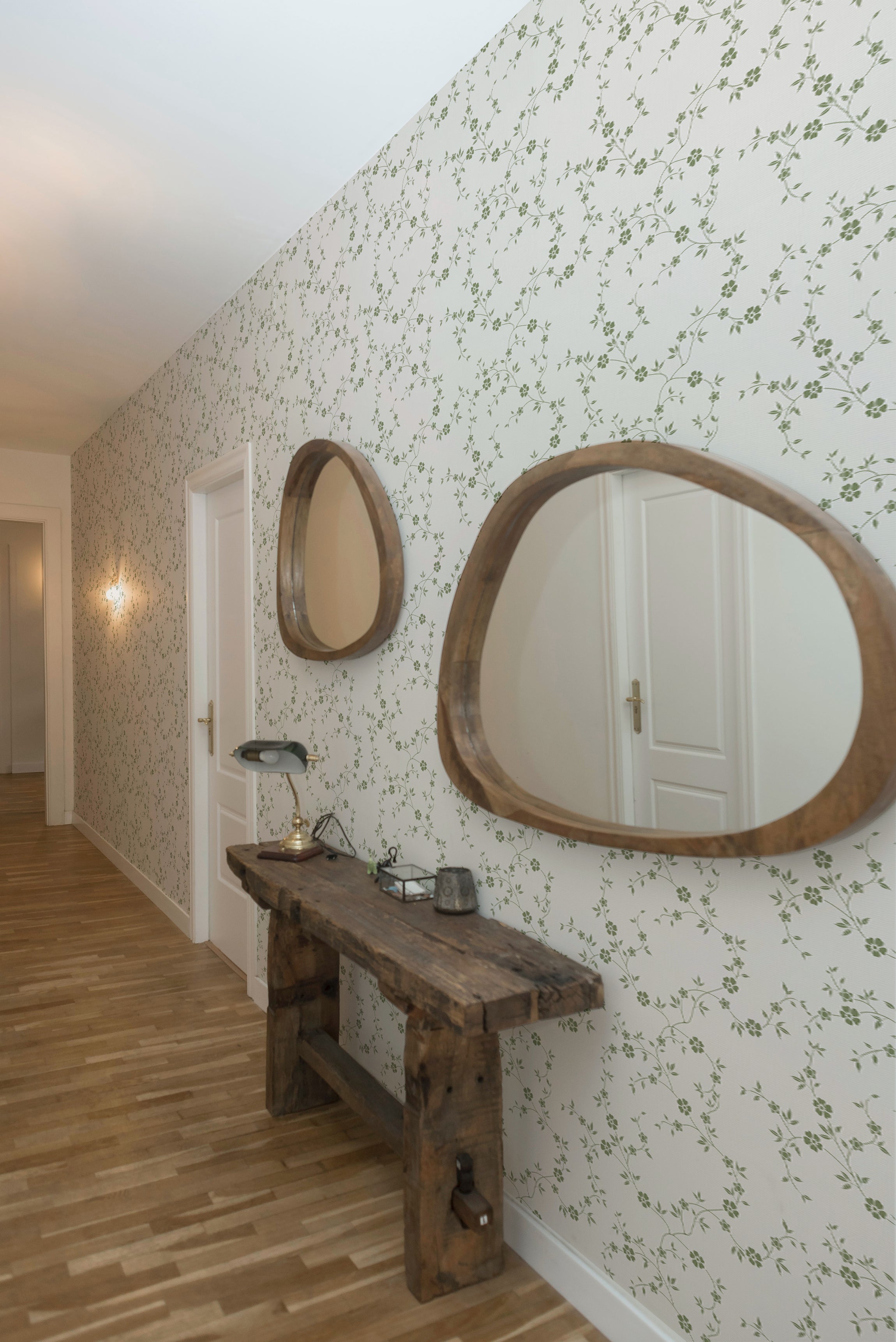 A spacious hallway is lined with the Charming Floral Wallpaper-Moss, enhancing the area with a classic, botanical charm. Two round wooden mirrors hang on the wallpapered wall above a rustic wooden bench, creating a welcoming and homey atmosphere.