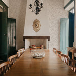 An elegant dining room with a large wooden table set against a wall adorned with 'Charming Floral Wallpaper', featuring a subtle pattern of beige flowers on an off-white background. A vintage chandelier hangs above, enhancing the room's classic charm.