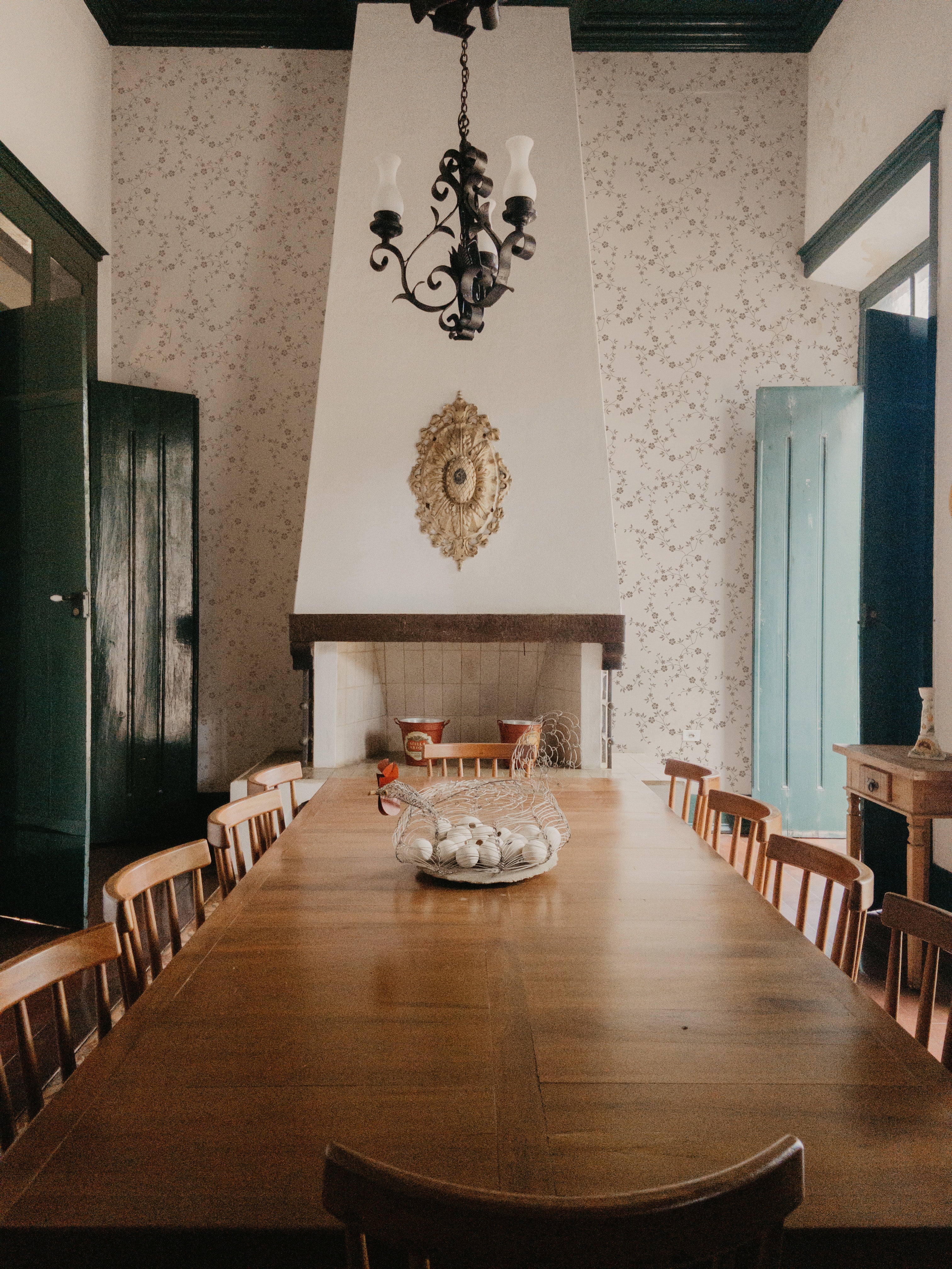 An elegant dining room with a large wooden table set against a wall adorned with 'Charming Floral Wallpaper', featuring a subtle pattern of beige flowers on an off-white background. A vintage chandelier hangs above, enhancing the room's classic charm.