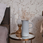 A cozy bedside with 'Charming Floral Wallpaper' providing a tranquil backdrop. The wallpaper's beige floral design exudes a peaceful atmosphere, paired with a nightstand that holds a blue vase and a brass lamp.