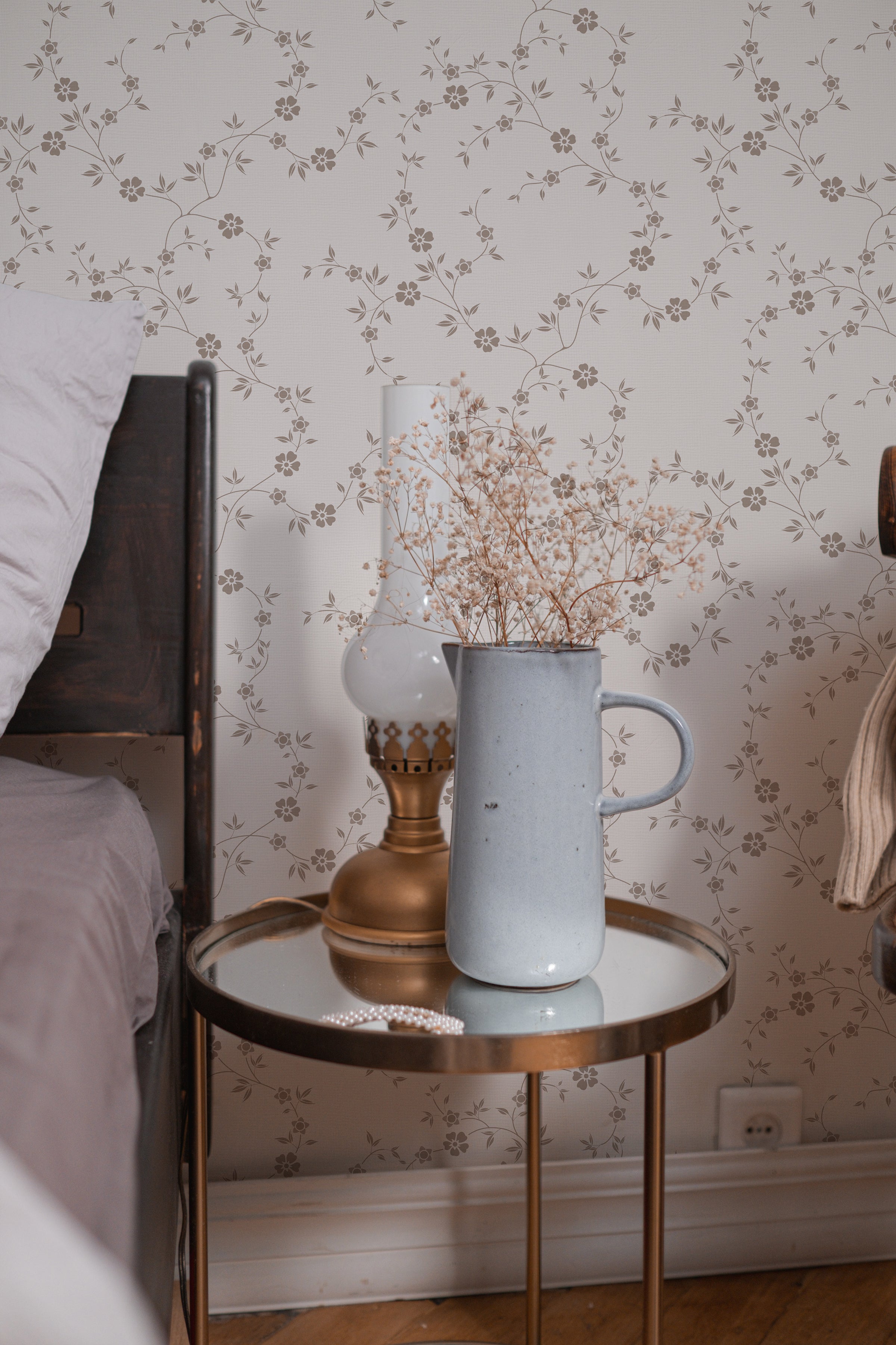 A cozy bedside with 'Charming Floral Wallpaper' providing a tranquil backdrop. The wallpaper's beige floral design exudes a peaceful atmosphere, paired with a nightstand that holds a blue vase and a brass lamp.