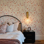 A beautifully appointed bedroom with a wall covered in Blushing Floral Wallpaper, featuring an array of muted pink, red, and purple flowers against a light cream background. The setting includes a bed with a dark metal frame, plush pillows, and a textured throw, complemented by a black nightstand with decorative items.