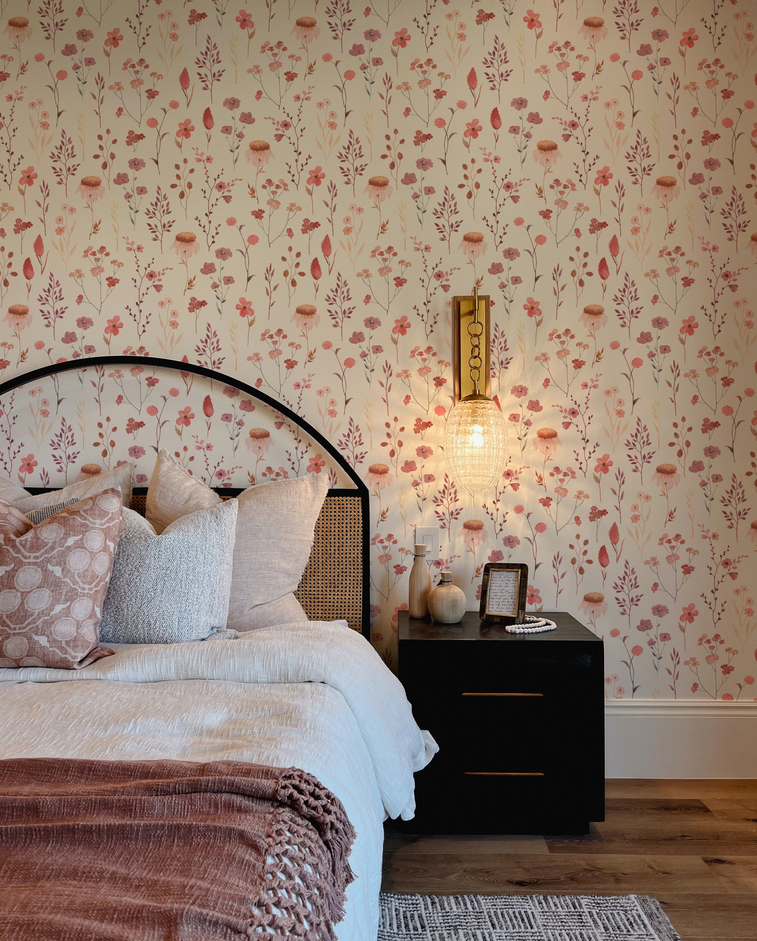A beautifully appointed bedroom with a wall covered in Blushing Floral Wallpaper, featuring an array of muted pink, red, and purple flowers against a light cream background. The setting includes a bed with a dark metal frame, plush pillows, and a textured throw, complemented by a black nightstand with decorative items.