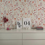 A stylish interior decor scene with Blushing Floral Wallpaper enhancing the wall behind a white modern dresser. The top of the dresser displays frames, books, and floral decorations that harmonize with the wallpaper's colors.