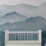 A serene nursery room featuring the Watercolour Mountains Wallpaper Mural, depicting soft watercolor mountains in shades of blue. The mural creates a tranquil backdrop behind a white crib, with plush toys and a small stool, adding a whimsical touch to the soothing space