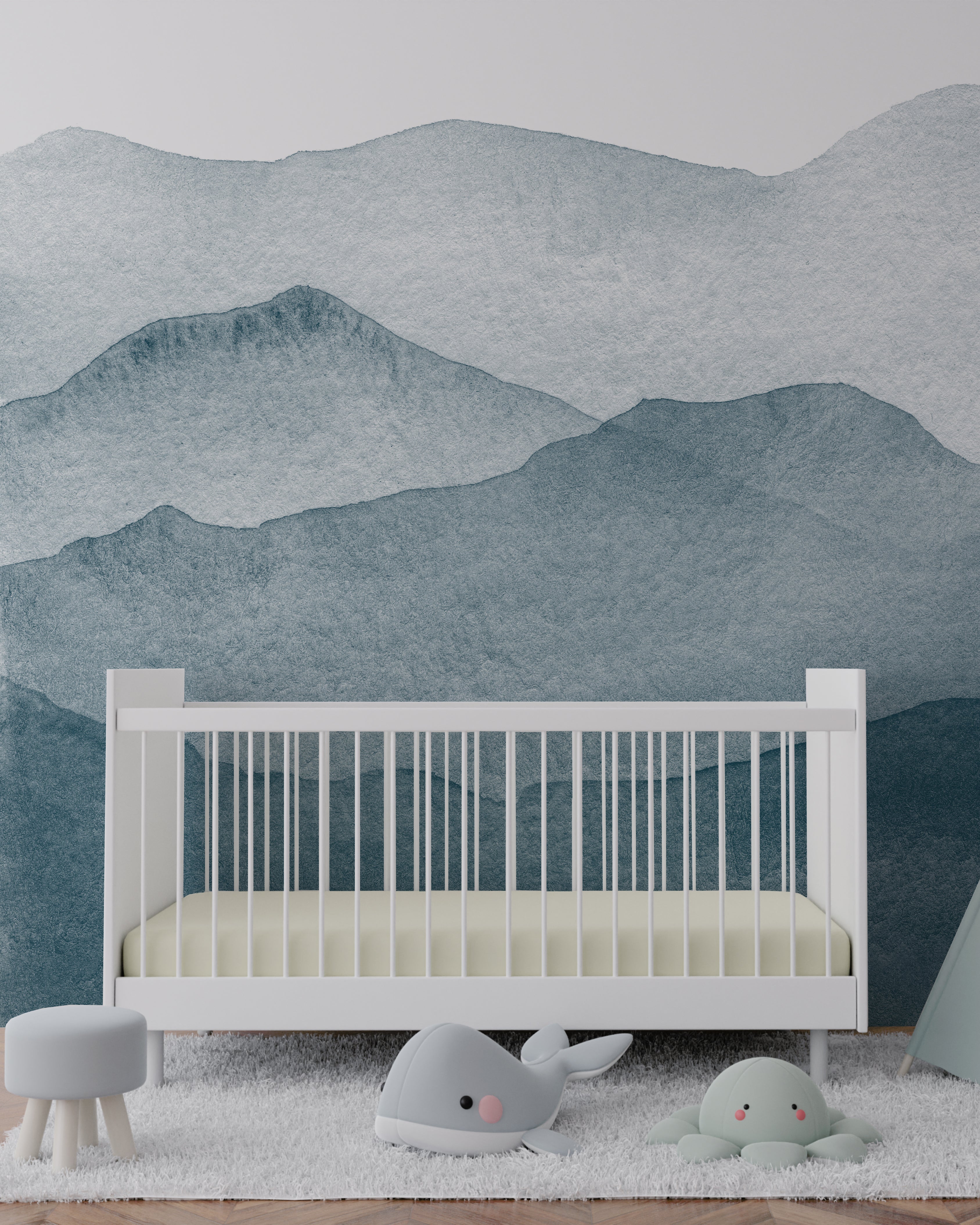A serene nursery room featuring the Watercolour Mountains Wallpaper Mural, depicting soft watercolor mountains in shades of blue. The mural creates a tranquil backdrop behind a white crib, with plush toys and a small stool, adding a whimsical touch to the soothing space
