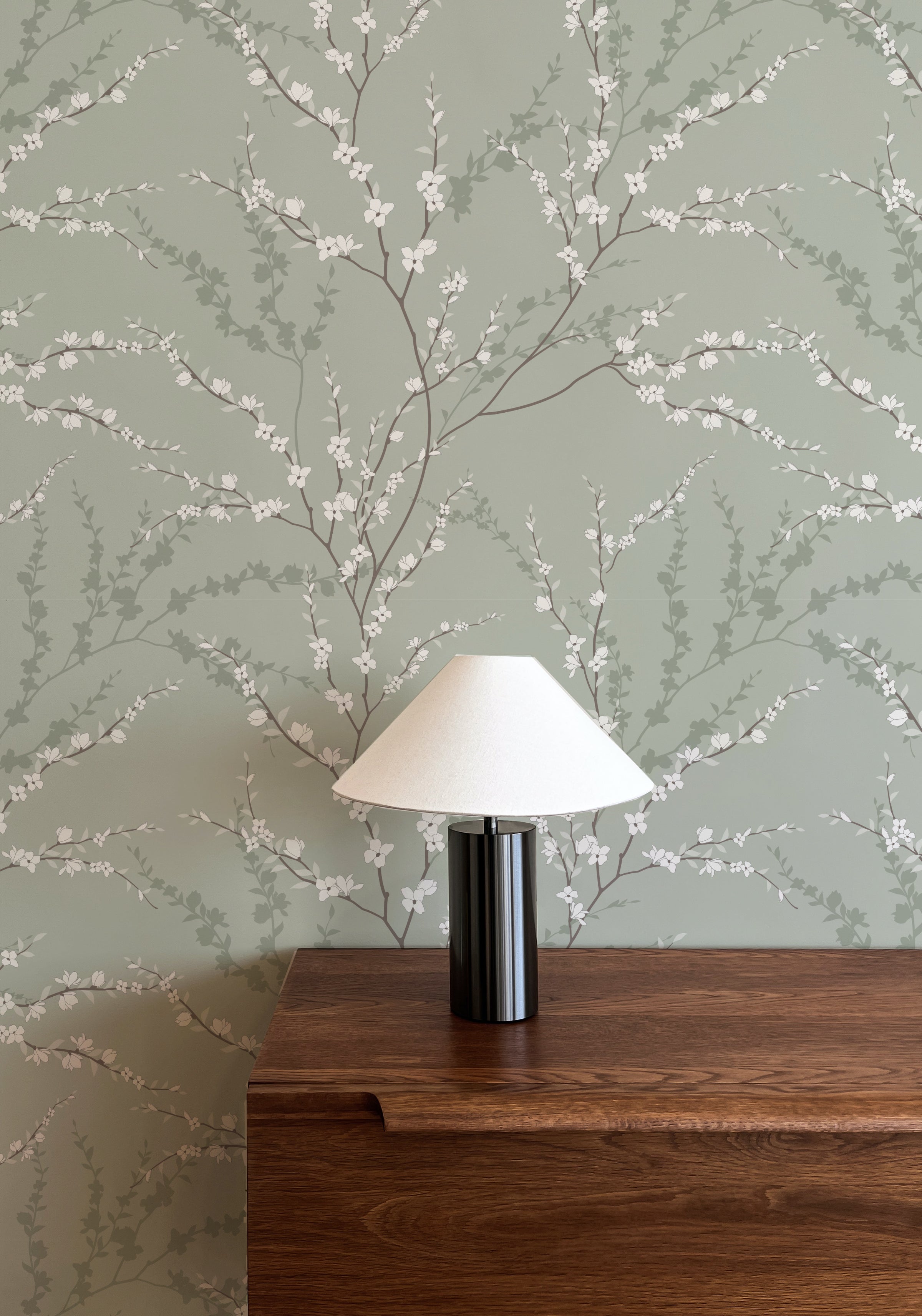 A chic table lamp stands against the Enchanted Blossoms Wallpaper, with its soft green hues and white flowers complementing the lamp's simple elegance, creating a harmonious blend of modern design and natural beauty.