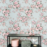 An elegant room with Rose Bouquet Wallpaper II showcasing a pattern of pink roses and green leaves on a light blue background. The decor includes a black shelving unit with candles, decorative boxes, and a bird ornament.