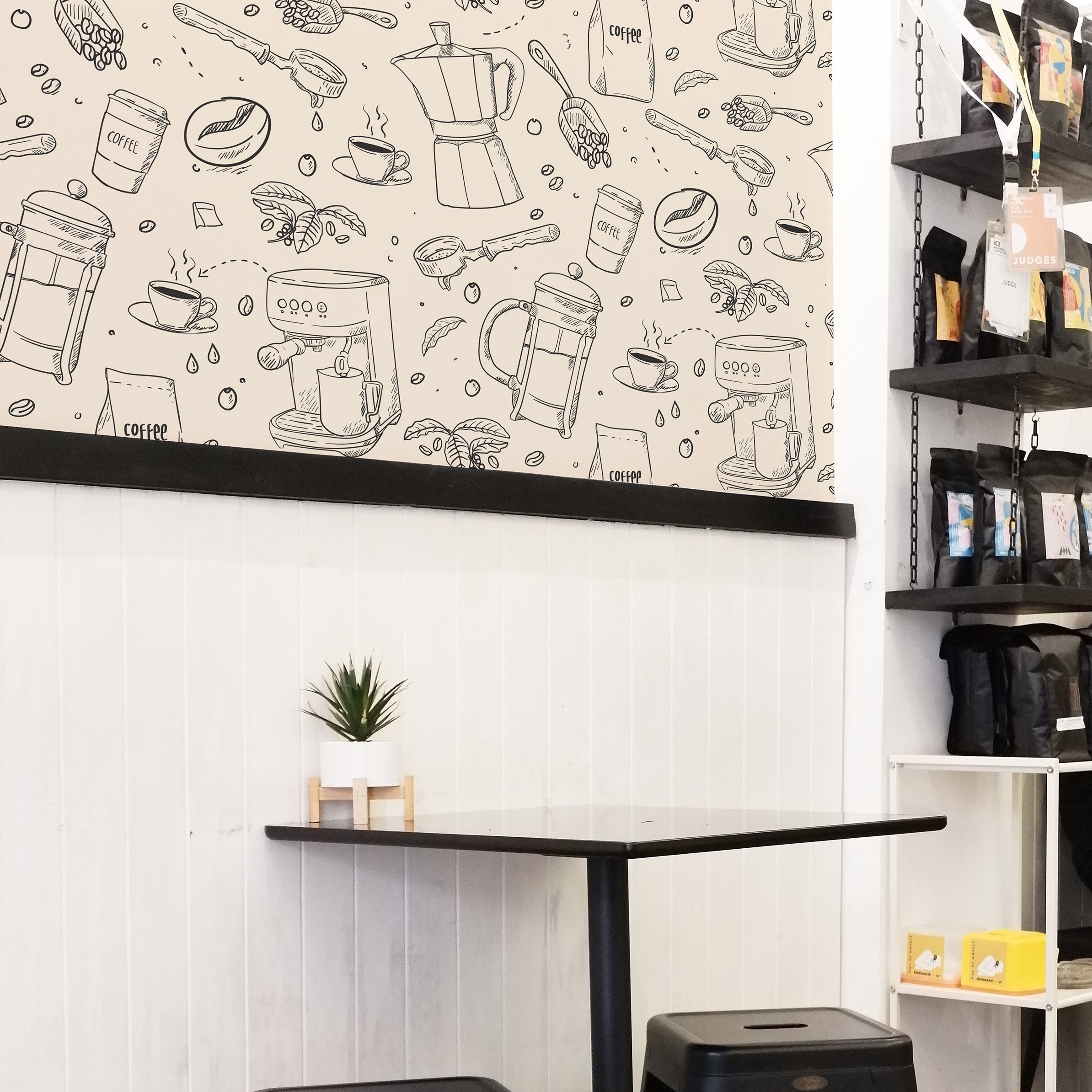 A cozy café corner featuring a unique wallpaper with hand-drawn illustrations of various coffee-making tools and beans. The wallpaper covers the upper half of the wall above a white subway-tiled lower half, accompanied by modern black metal stools and a minimalist table.