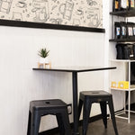 A cozy café corner featuring a unique wallpaper with hand-drawn illustrations of various coffee-making tools and beans. The wallpaper covers the upper half of the wall above a white subway-tiled lower half, accompanied by modern black metal stools and a minimalist table.