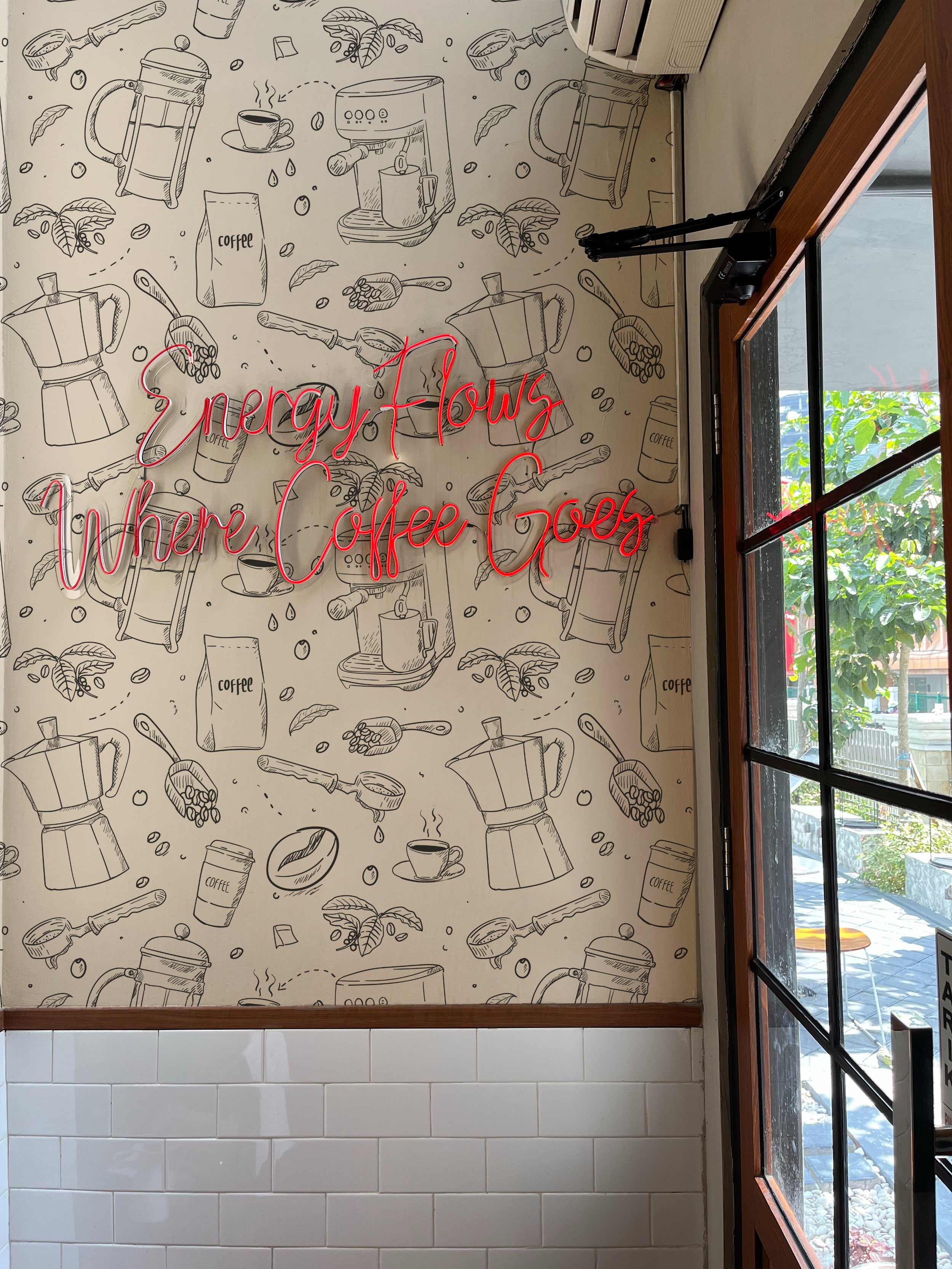 A vibrant wall mural in a café displaying a playful and detailed drawing of coffee accessories like grinders, cups, and beans. A neon sign stating "Energy Flows Where Coffee Goes" adds a striking contrast against the sketch-style wallpaper.