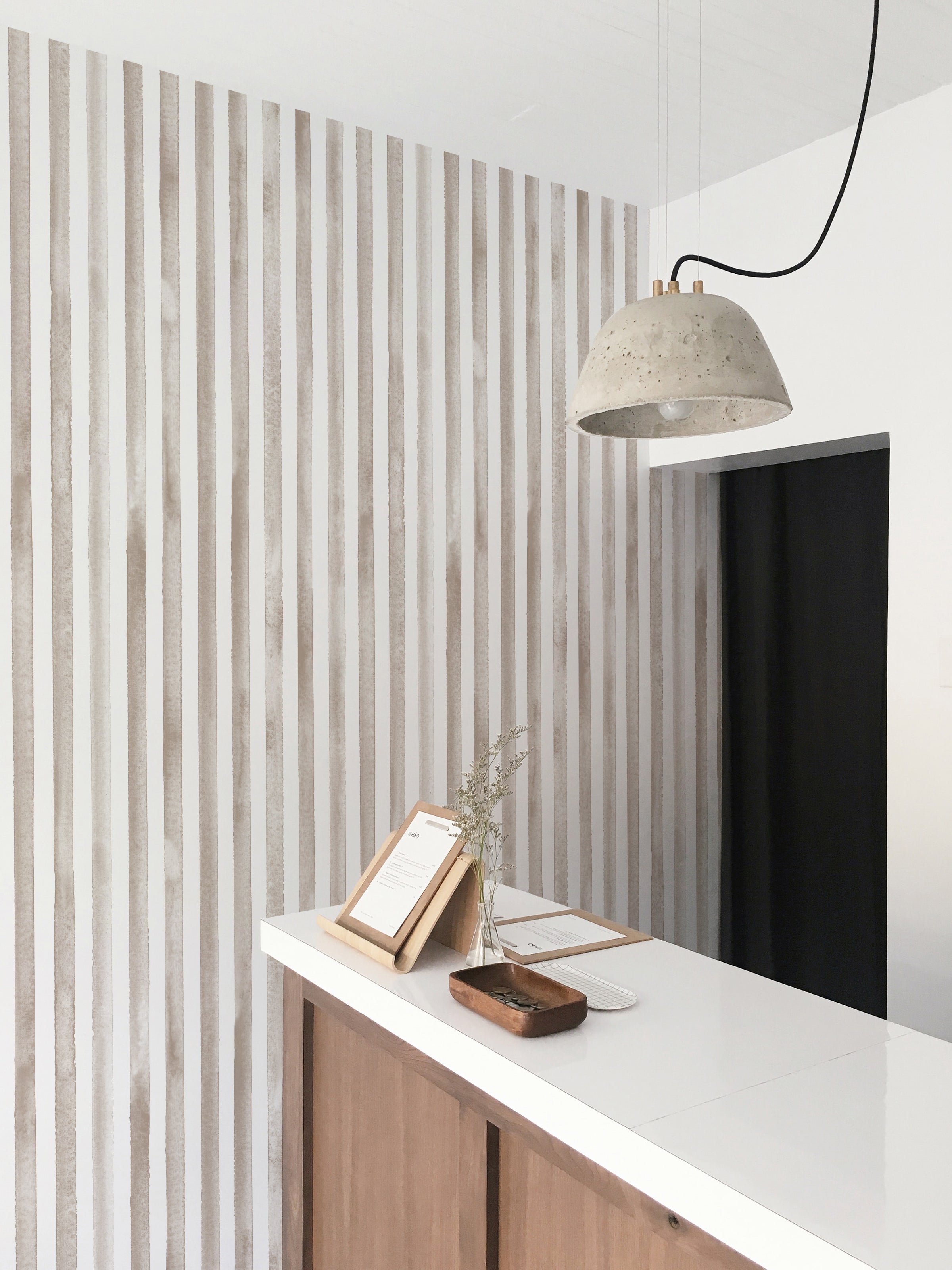A stylish home office setup with a white countertop against a background of vertical watercolour stripes wallpaper. The decor includes a modern chair, a concrete pendant lamp, and minimalistic accessories, emphasizing a clean and contemporary workspace.
