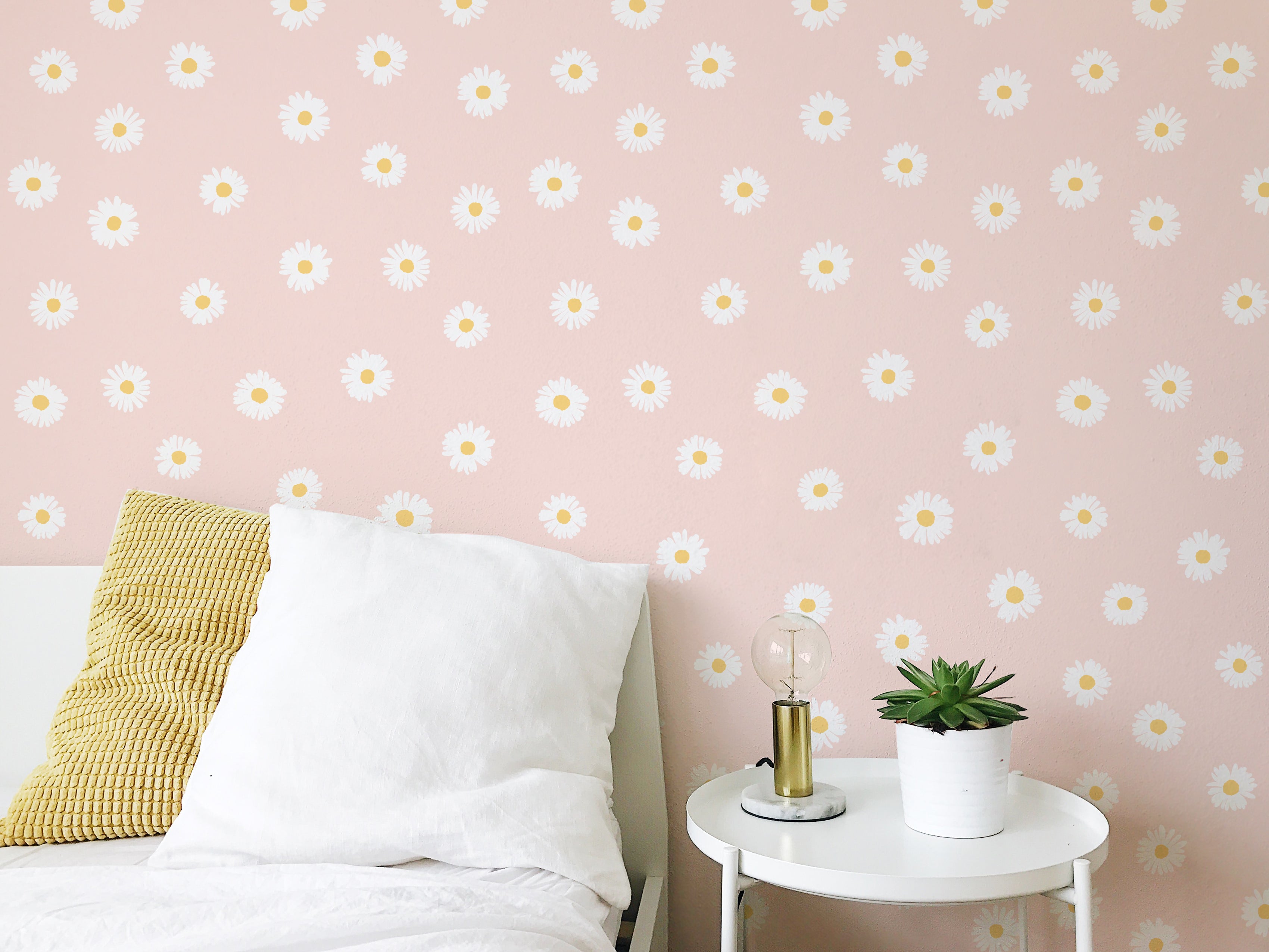 A cozy bedroom corner featuring a pastel pink wall adorned with Daisy Daze Wallpaper. The wallpaper has a light peach background with a pattern of white daisies with yellow centers. Next to the bed, covered with white linens, sits a small round table with a lamp and a potted plant, enhancing the room's cheerful and airy feel.